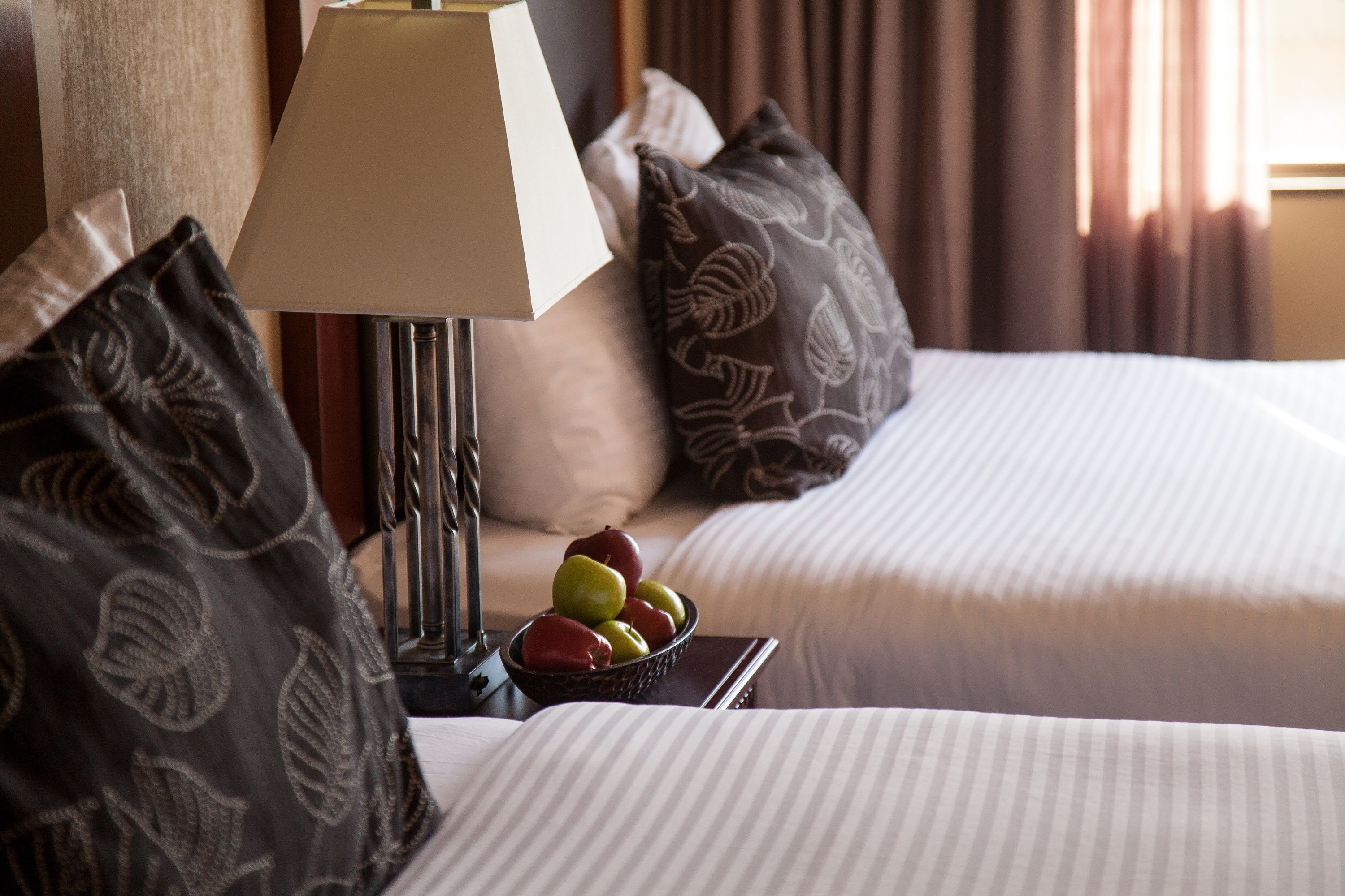 Habitación de hotel con dos camas, una mesita de noche con lámpara, un frutero y almohadas estampadas.