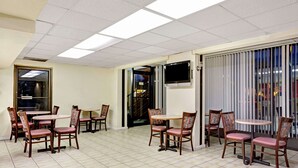 A hotel lobby with multiple dining tables and chairs, a flat-screen TV mounted on the wall, and a large window with blinds.