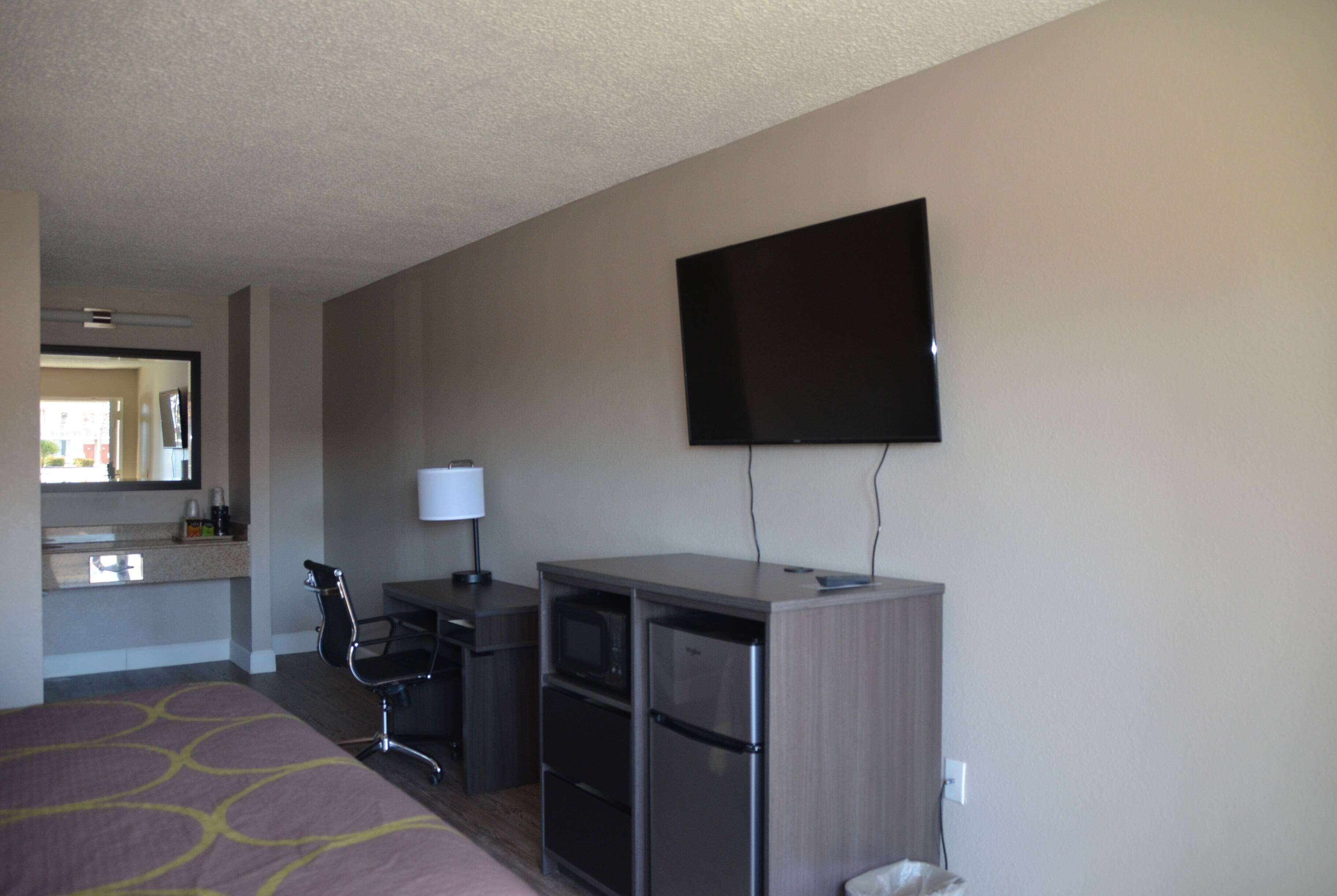 A hotel room with a bed, desk, chair, microwave, refrigerator, and a flat-screen TV mounted on the wall.