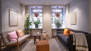 A living room with a sofa, ottoman, and a coffee table. There are two windows with plants and pendant lights. - Hotel Mayfair (Copenhagen)