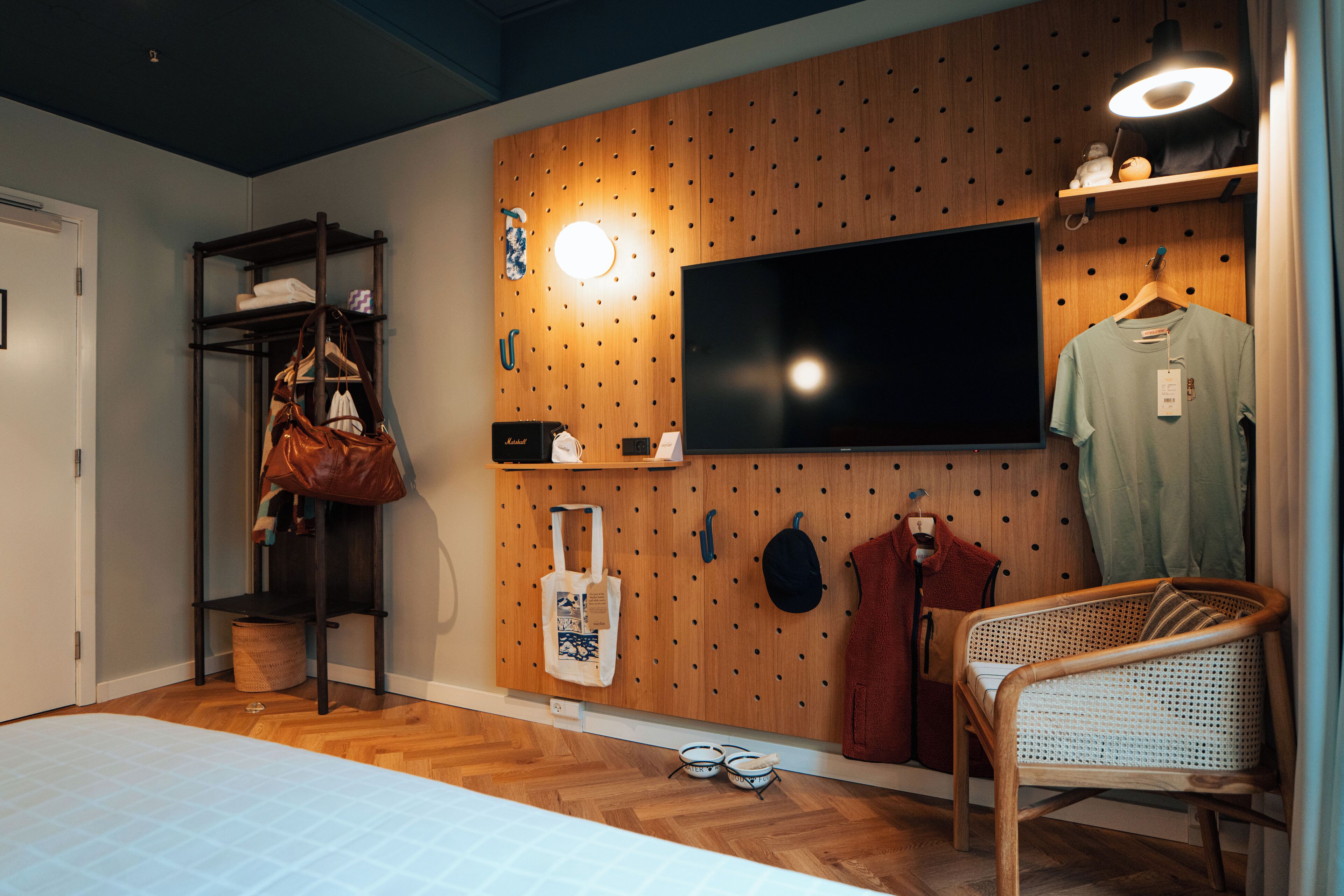 Ein Zimmer mit einer Holzvertäfelung, einem Fernseher über einem Kleiderständer, einem Korbstuhl und einem Bett mit gemustertem Überwurf.