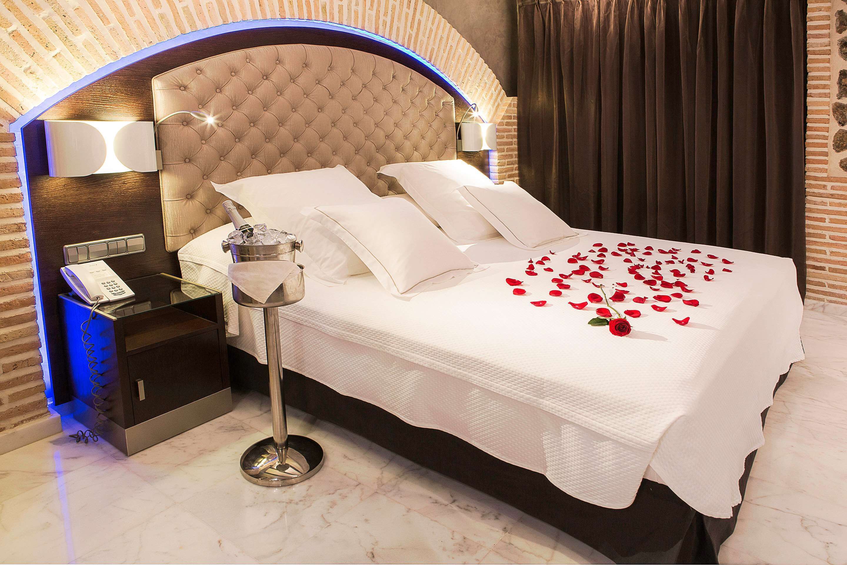 Una cama bien hecha con sábanas blancas y pétalos de rosa, un teléfono en la mesita de noche y un minibar.