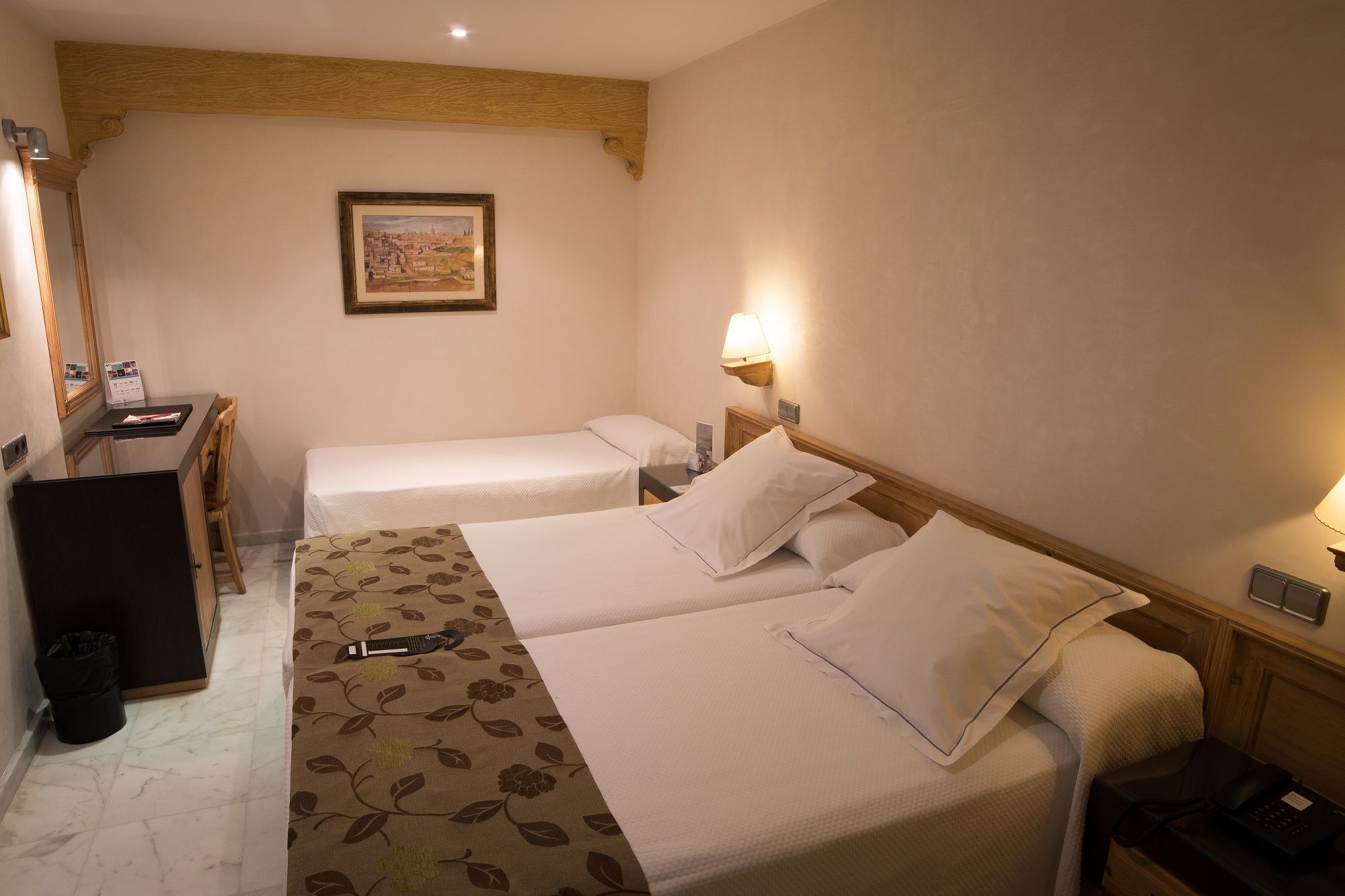 Habitación de hotel con dos camas individuales, un escritorio con silla, un cubo de basura pequeño y un cuadro en la pared.