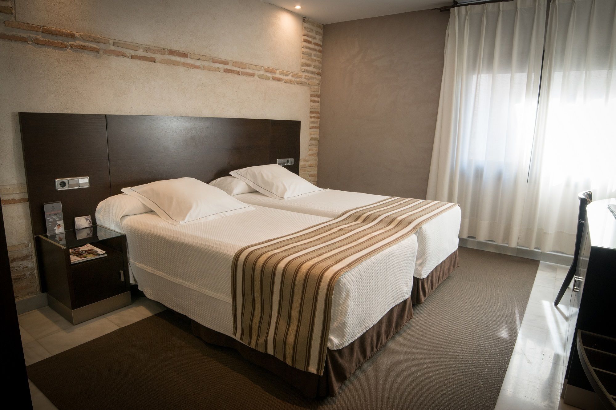 A double bed with a striped blanket, a nightstand with a lamp, and a brick wall.