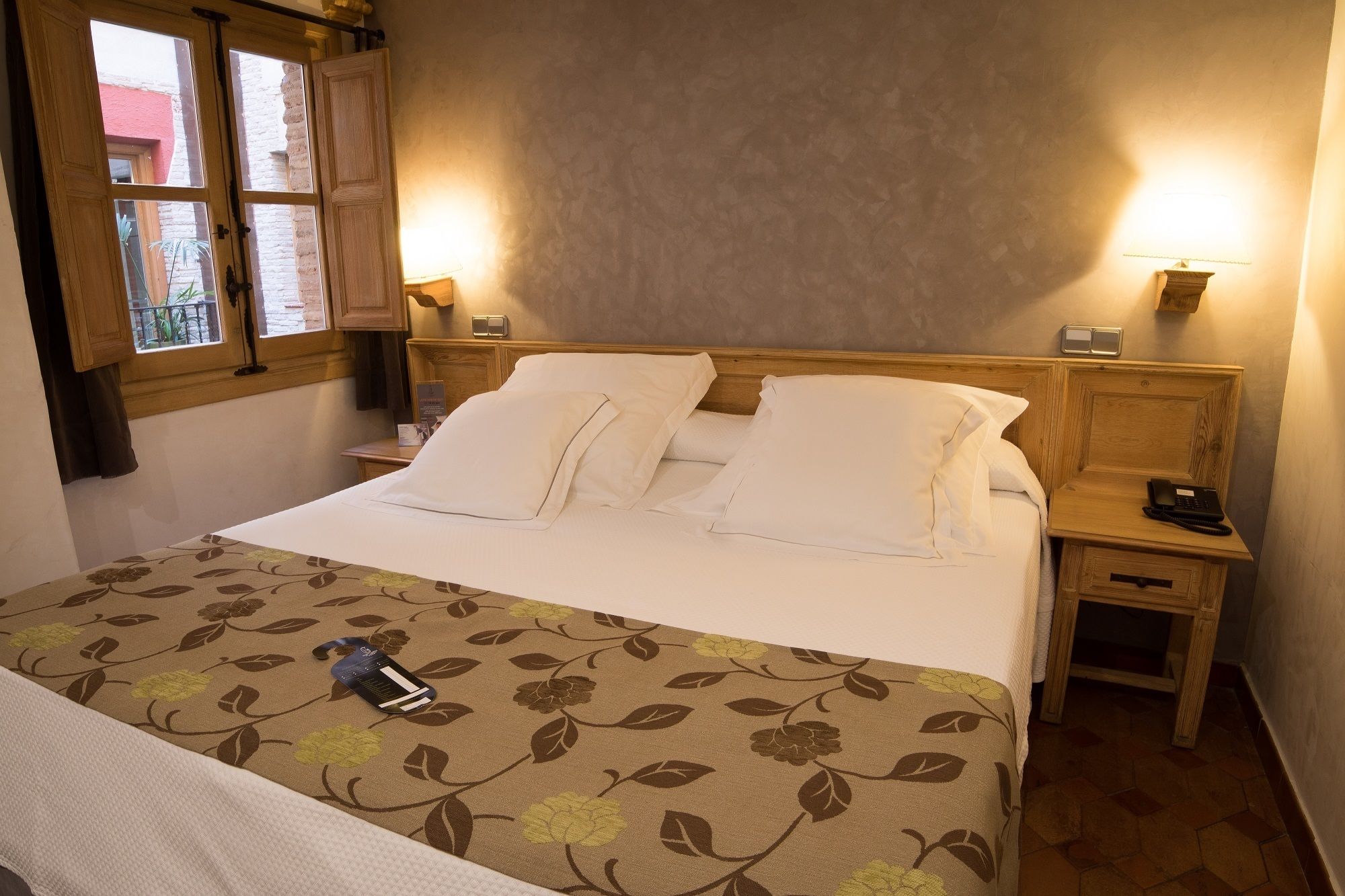 Ein ordentlich bezogenes Bett mit weißer Bettwäsche, einem floralen Überwurf und einem Nachttisch mit Lampe und Telefon.