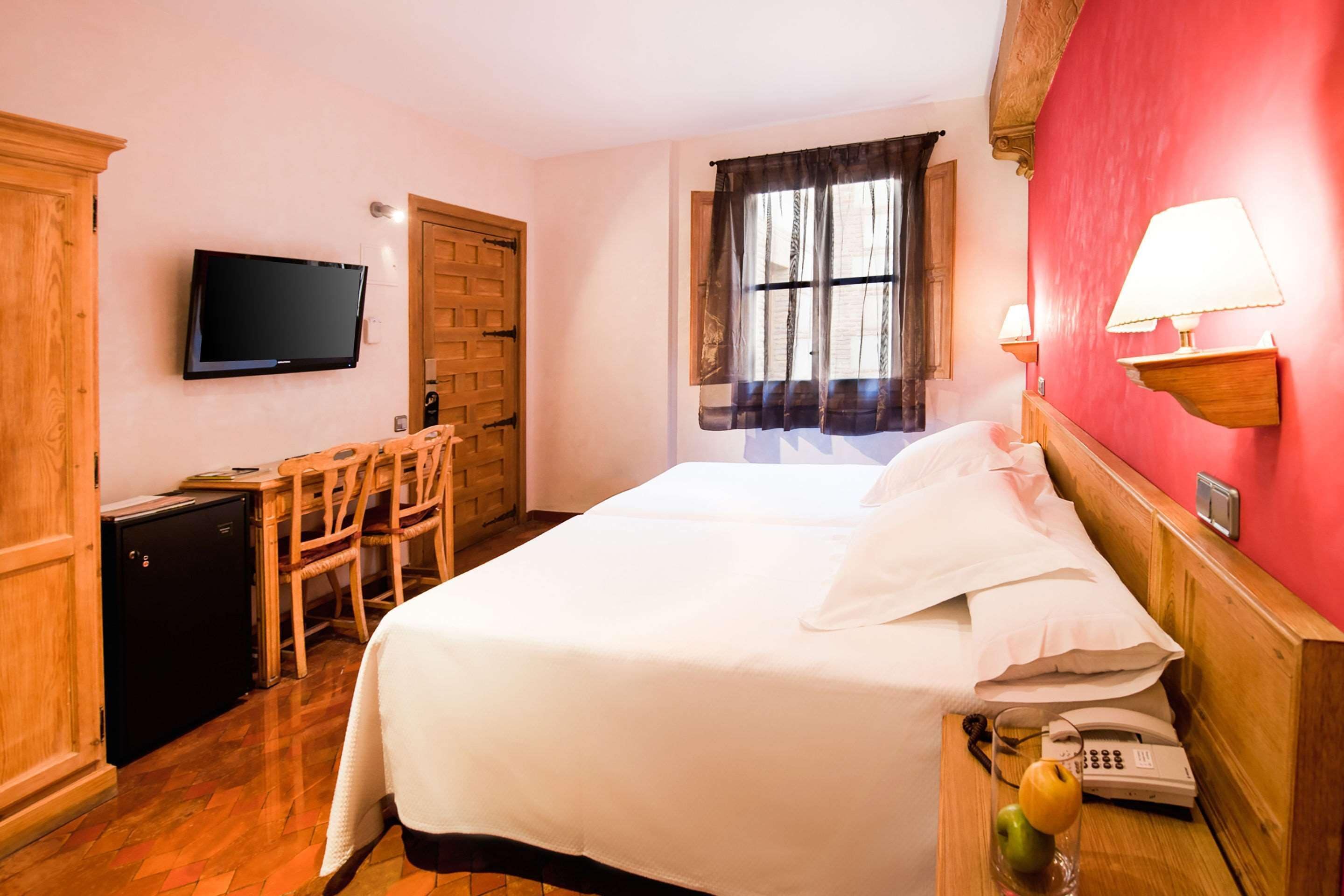 A hotel room with a bed, a desk, a television, a window with curtains, and a wall-mounted lamp.