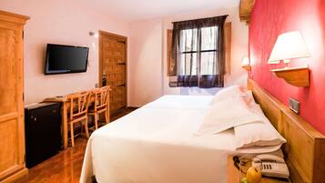 A hotel room with a bed, a desk, a television, a window with curtains, and a wall-mounted lamp.