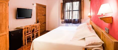A hotel room with a bed, a desk, a television, a window with curtains, and a wall-mounted lamp.