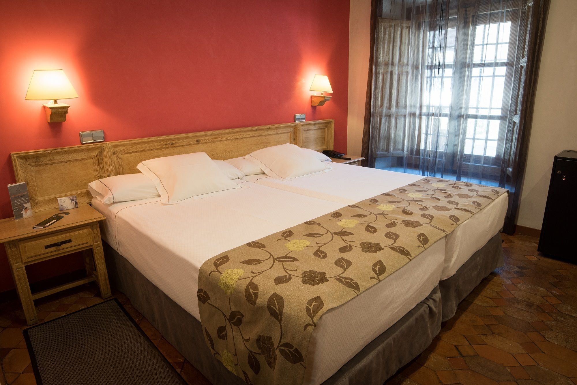 Una cama doble con colcha estampada, dos lámparas de noche, un cabecero de madera, una mesita de noche con un teléfono y un folleto, y una ventana con cortinas traslúcidas.