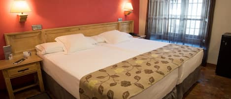 Una cama doble con colcha estampada, dos lámparas de noche, un cabecero de madera, una mesita de noche con un teléfono y un folleto, y una ventana con cortinas traslúcidas.