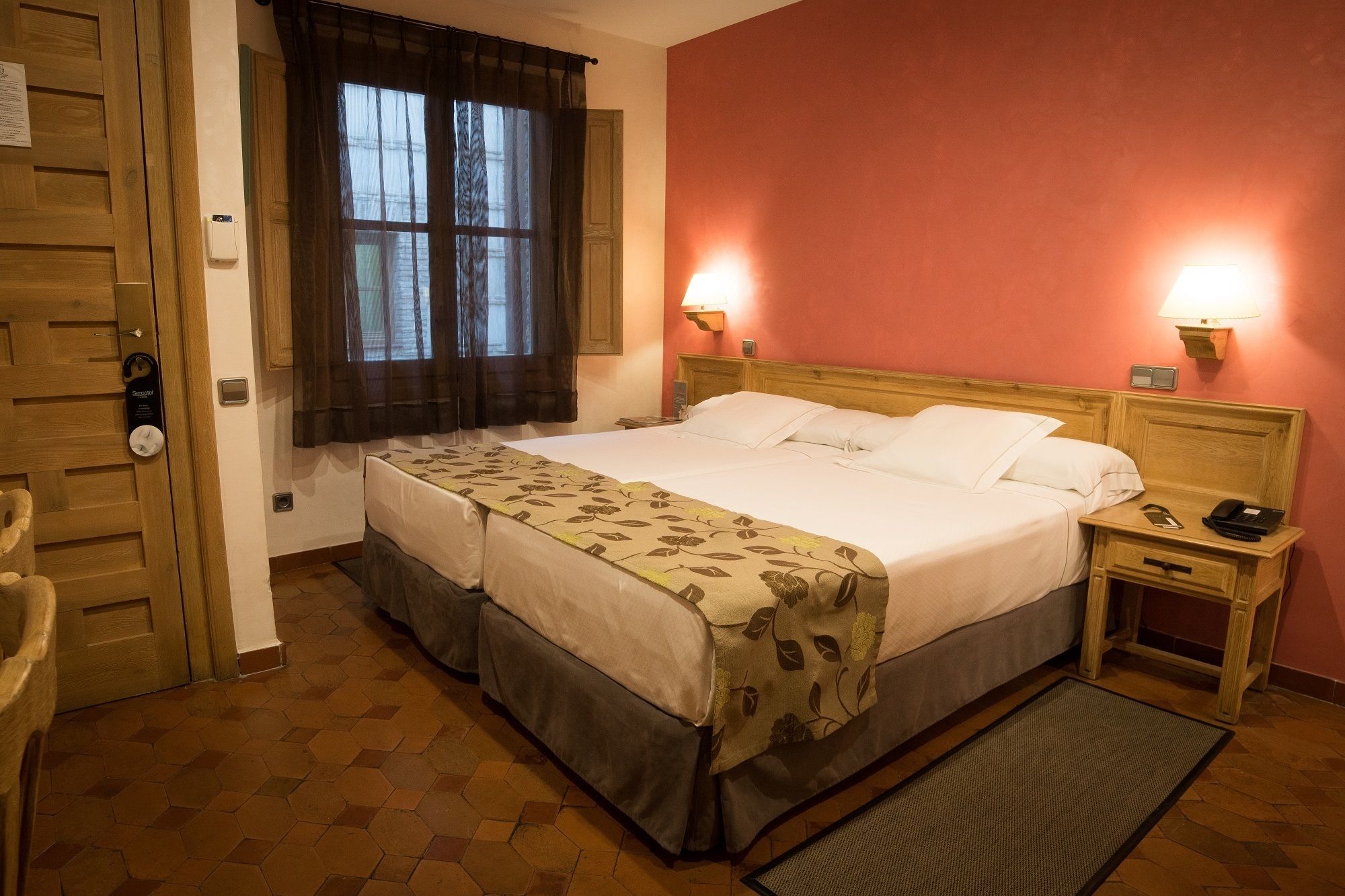 Une chambre d’hôtel avec un lit double, deux tables de chevet, une petite table, une fenêtre avec des rideaux, une porte avec une serrure et une couvre-lit à motifs.