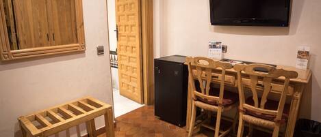A compact room with a wooden cabinet, a small black refrigerator, a television mounted on the wall, and a seating area with two wooden chairs and a small table.