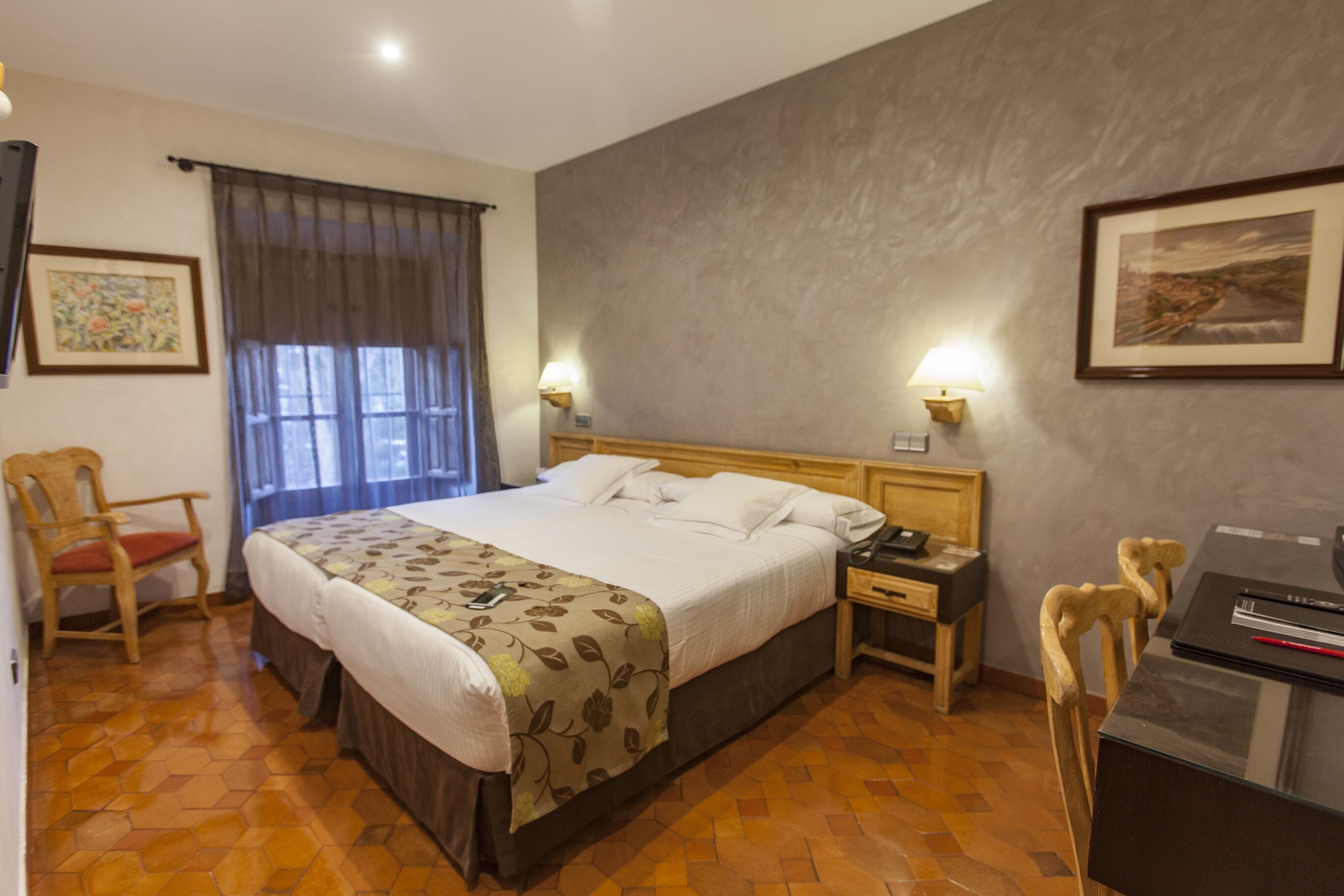 Habitación de hotel con una cama grande, cabecera de madera, dos mesitas de noche, un escritorio con una silla, una ventana con cortinas y una lámpara de pared.