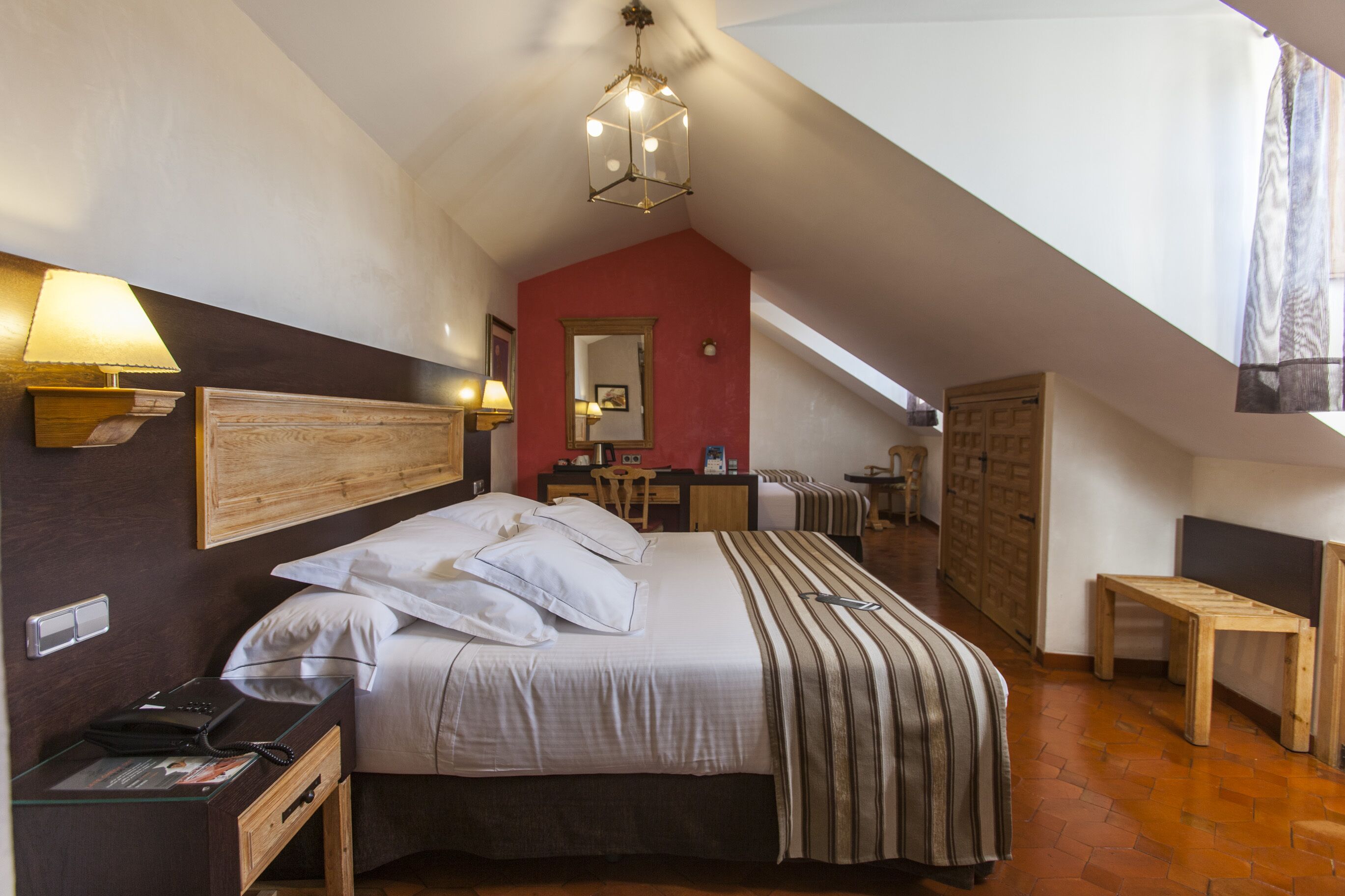 A bedroom with a bed, bedside lamps, a wooden headboard, a red accent wall, a mirror, a desk, a chair, a wooden bench, and a window with curtains.