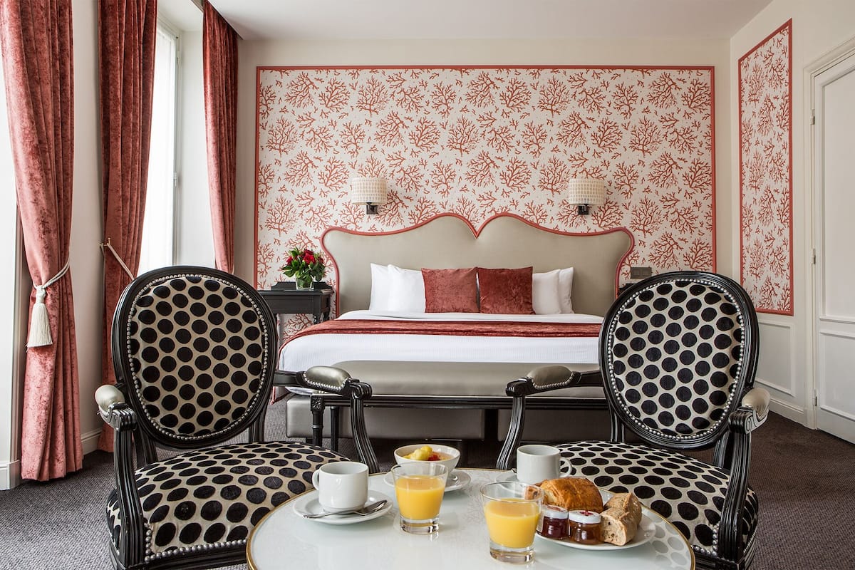 Habitación de hotel con cama, dos sillas, una mesa con desayuno y un papel tapiz estampado.