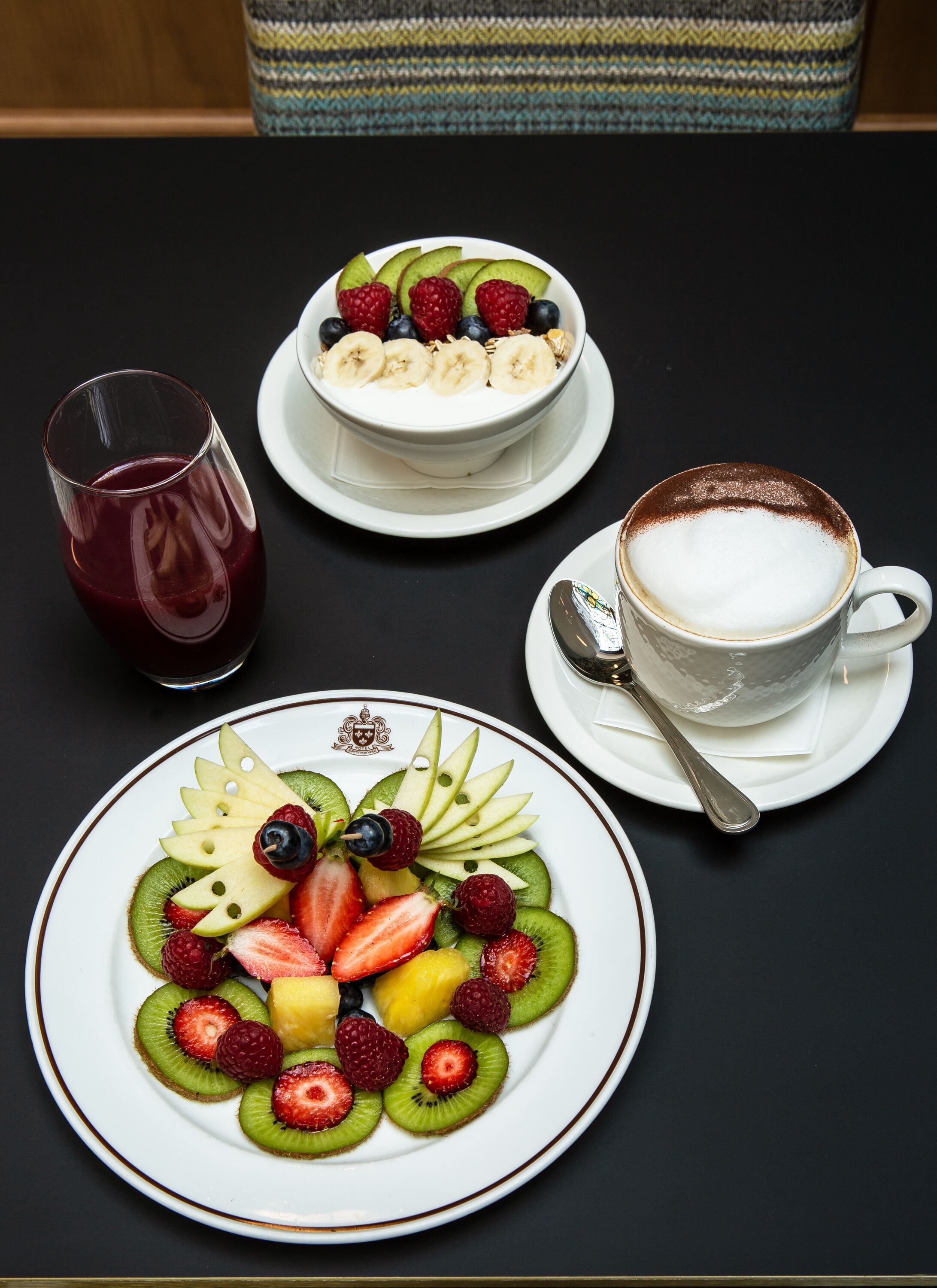 A table with a fruit platter, a bowl of yogurt with fruits, a glass of juice, and a cup of coffee.