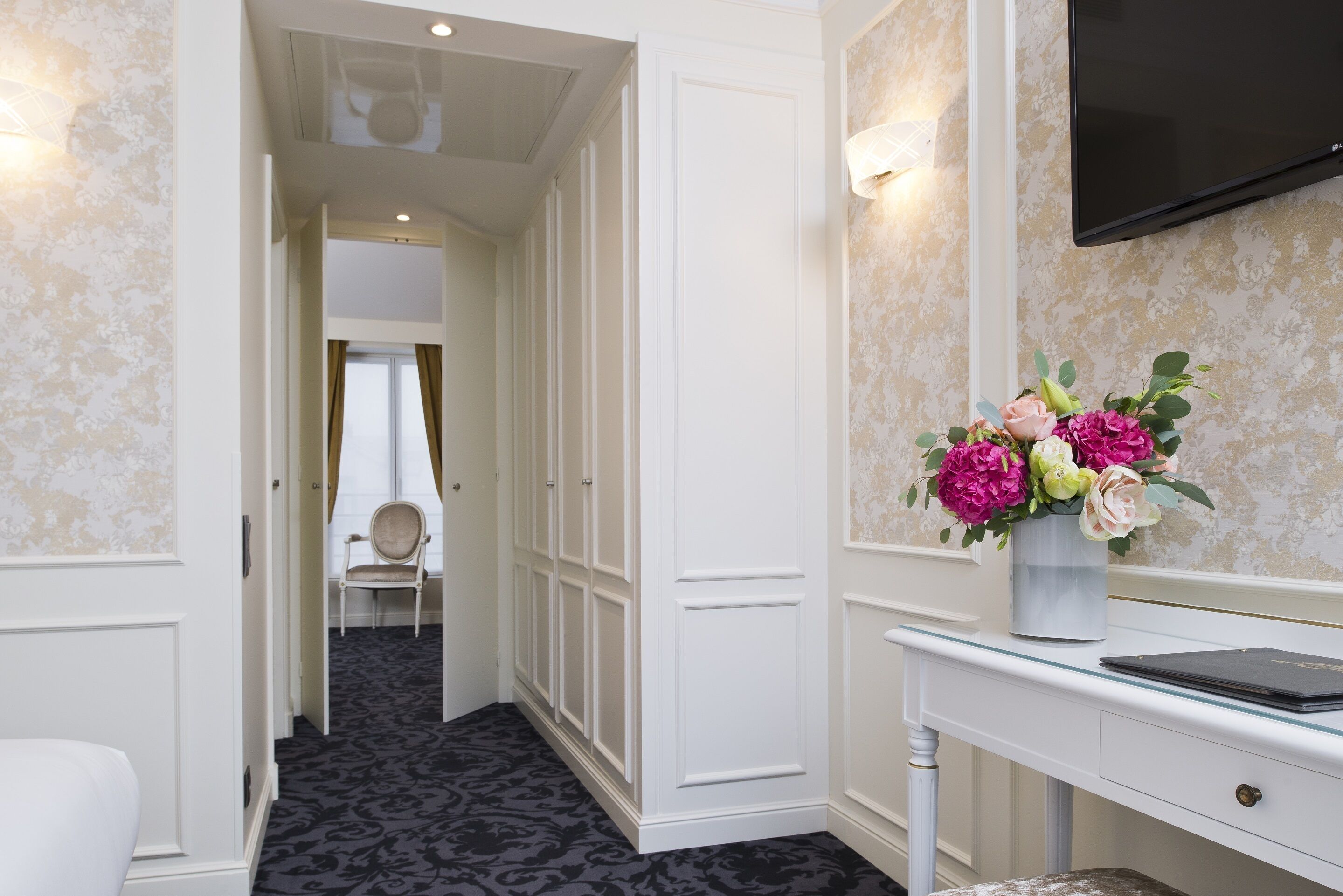 Habitación de hotel con alfombra de diseño oscuro, cómoda blanca con un florero y un televisor montado en la pared.