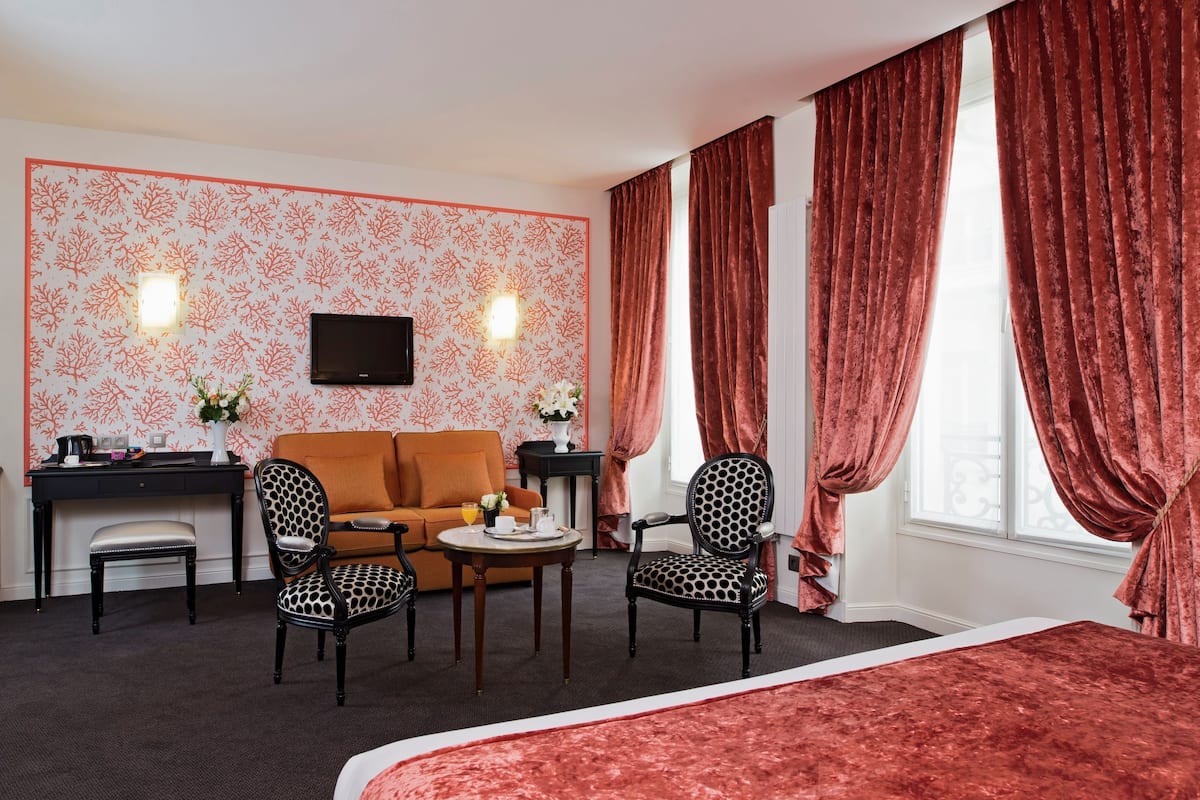 Habitación de hotel con sofá, dos sillones, una mesita con un florero y flores, un escritorio con televisor de pantalla plana y ventanas amplias con cortinas.
