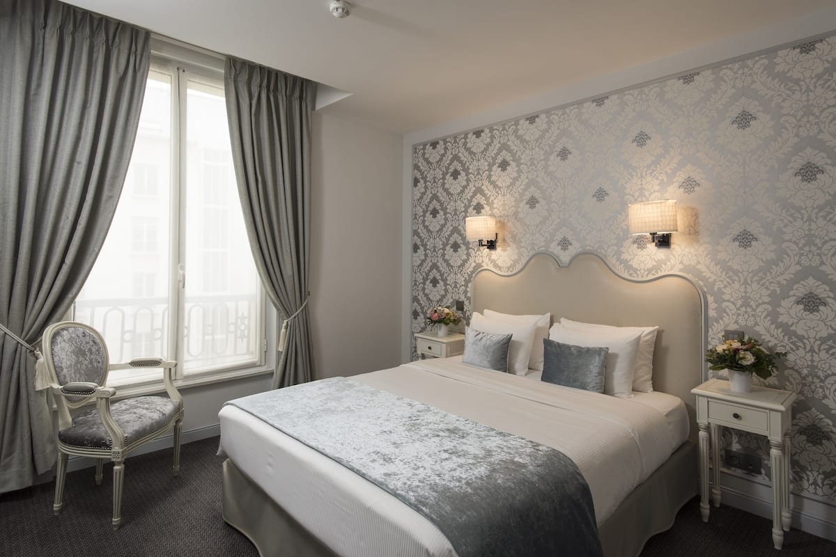 Una habitación de hotel con una cama grande, dos mesitas de noche con lámparas, una silla y una ventana con cortinas.