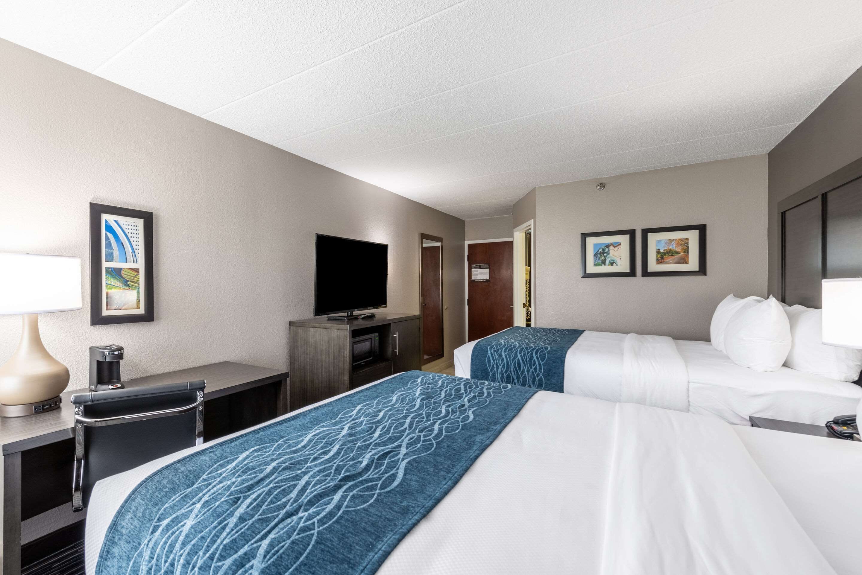 A hotel room with two beds, a desk with a lamp, a TV, and framed pictures on the wall.