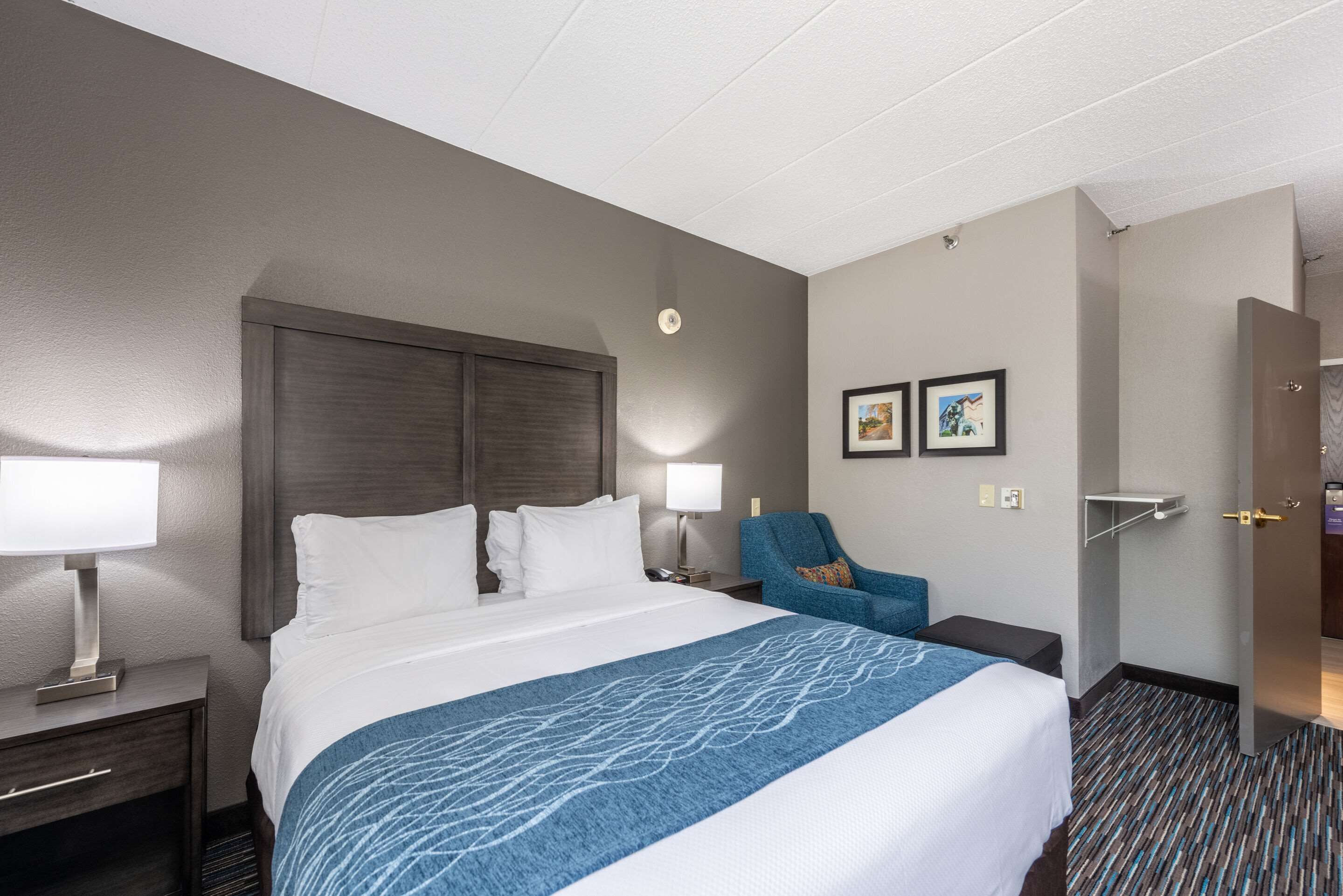 Habitación de hotel con una cama grande, una mesita de noche, una lámpara de escritorio, una silla y cuadros en la pared.