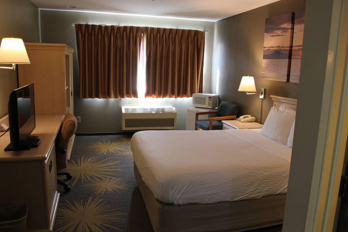 A hotel room with a bed, a desk with a lamp, a television, an air conditioning unit, a telephone, and a window with curtains.