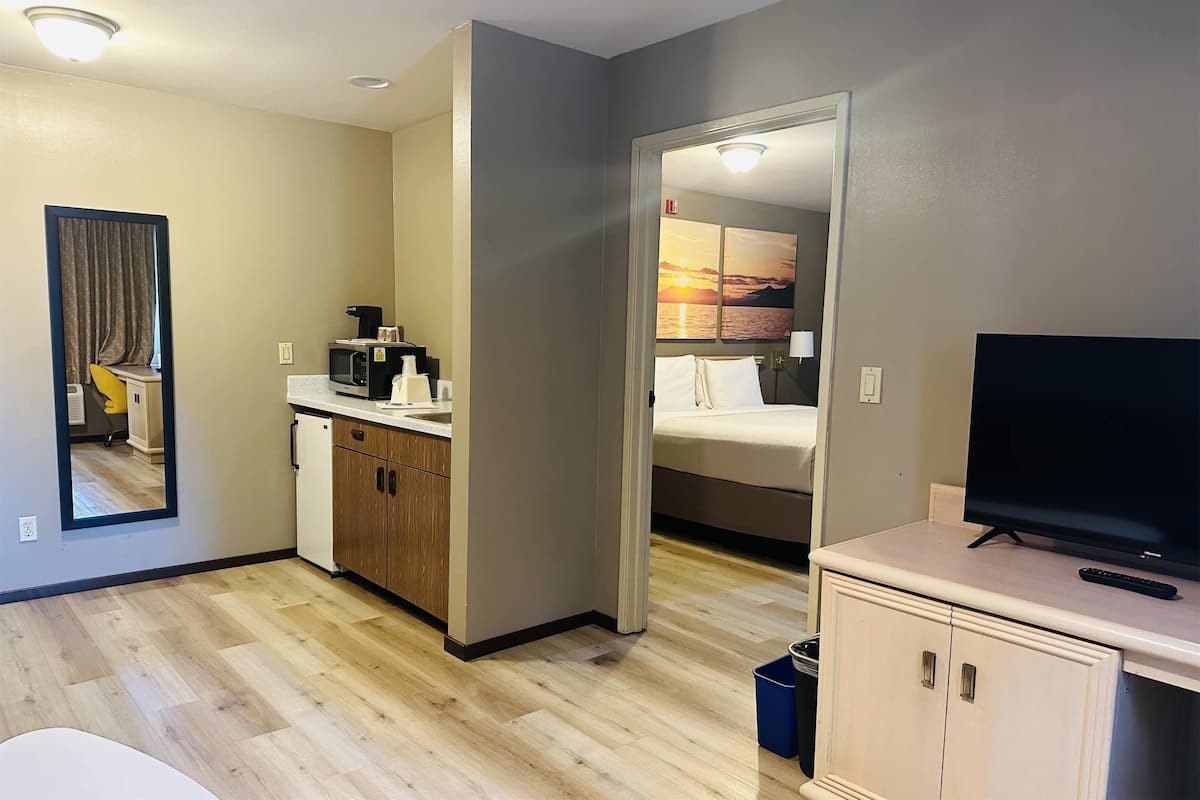 A compact hotel room with a kitchenette, a bed with a view of the sunset, a television, and a mirror.