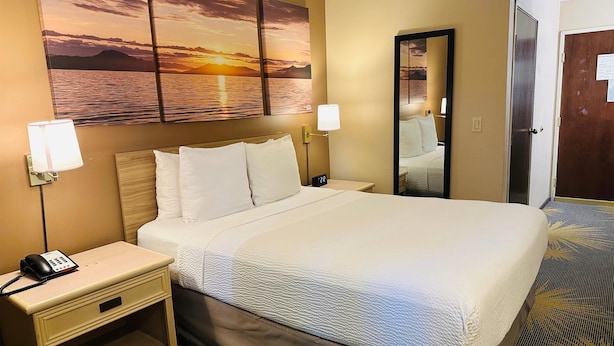 A hotel room with a large bed, two bedside lamps, a nightstand with a phone, a wall-mounted mirror, and a sunset painting above the bed. - Days Inn by Wyndham Seatac Airport (SeaTac)