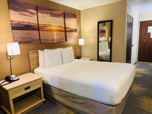 A hotel room with a large bed, two bedside lamps, a nightstand with a phone, a wall-mounted mirror, and a sunset painting above the bed. - Days Inn by Wyndham Seatac Airport (SeaTac)