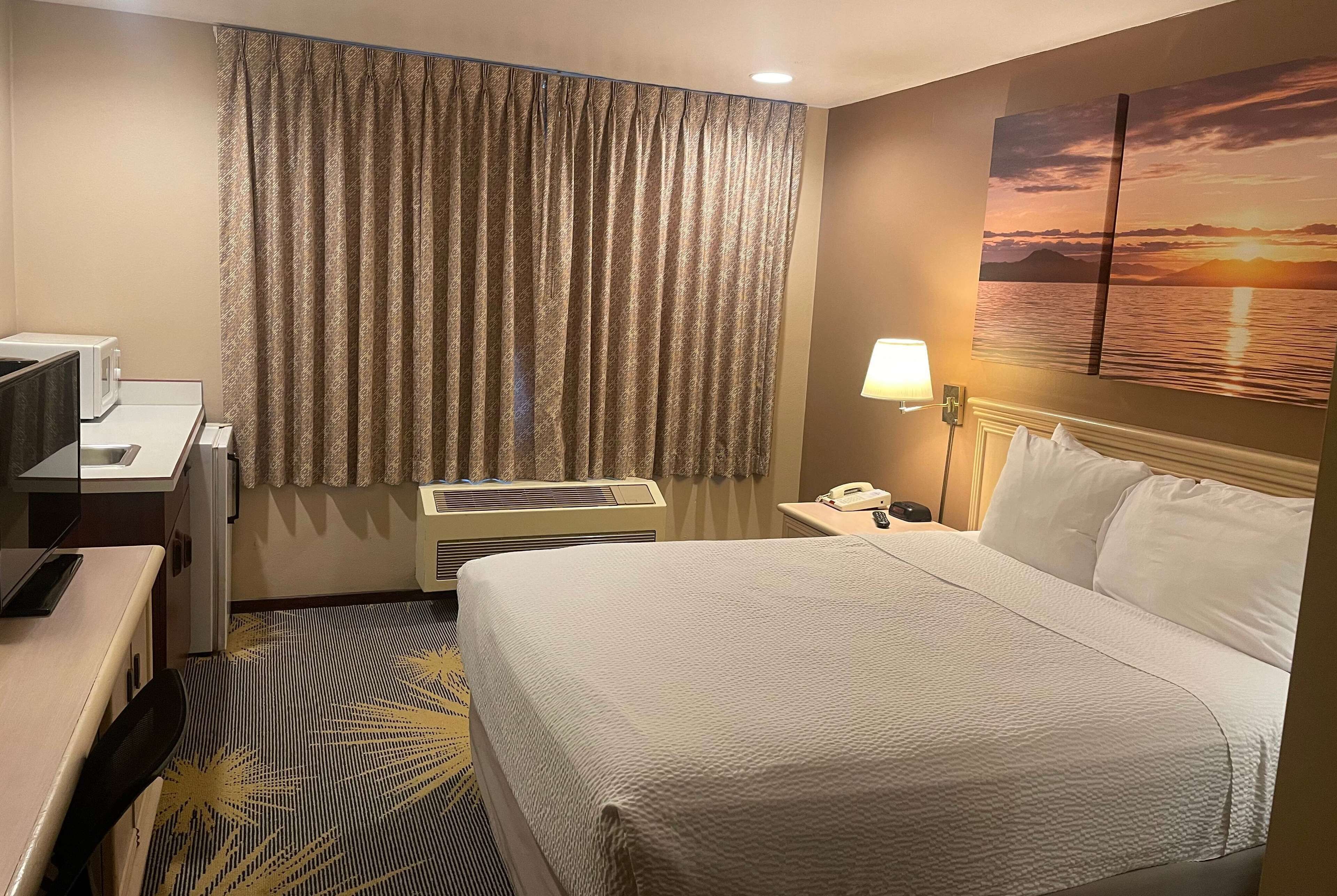 Une chambre d’hôtel avec un lit, un bureau équipé d’un ordinateur, un climatiseur, une table de chevet avec une lampe et un tableau représentant un coucher de soleil sur l’eau, accroché au mur.