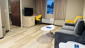 A modern living room with a sofa, a small table, a desk with a chair, and a television. There is a window with curtains, a door, and a radiator. - Days Inn by Wyndham Seatac Airport (SeaTac)