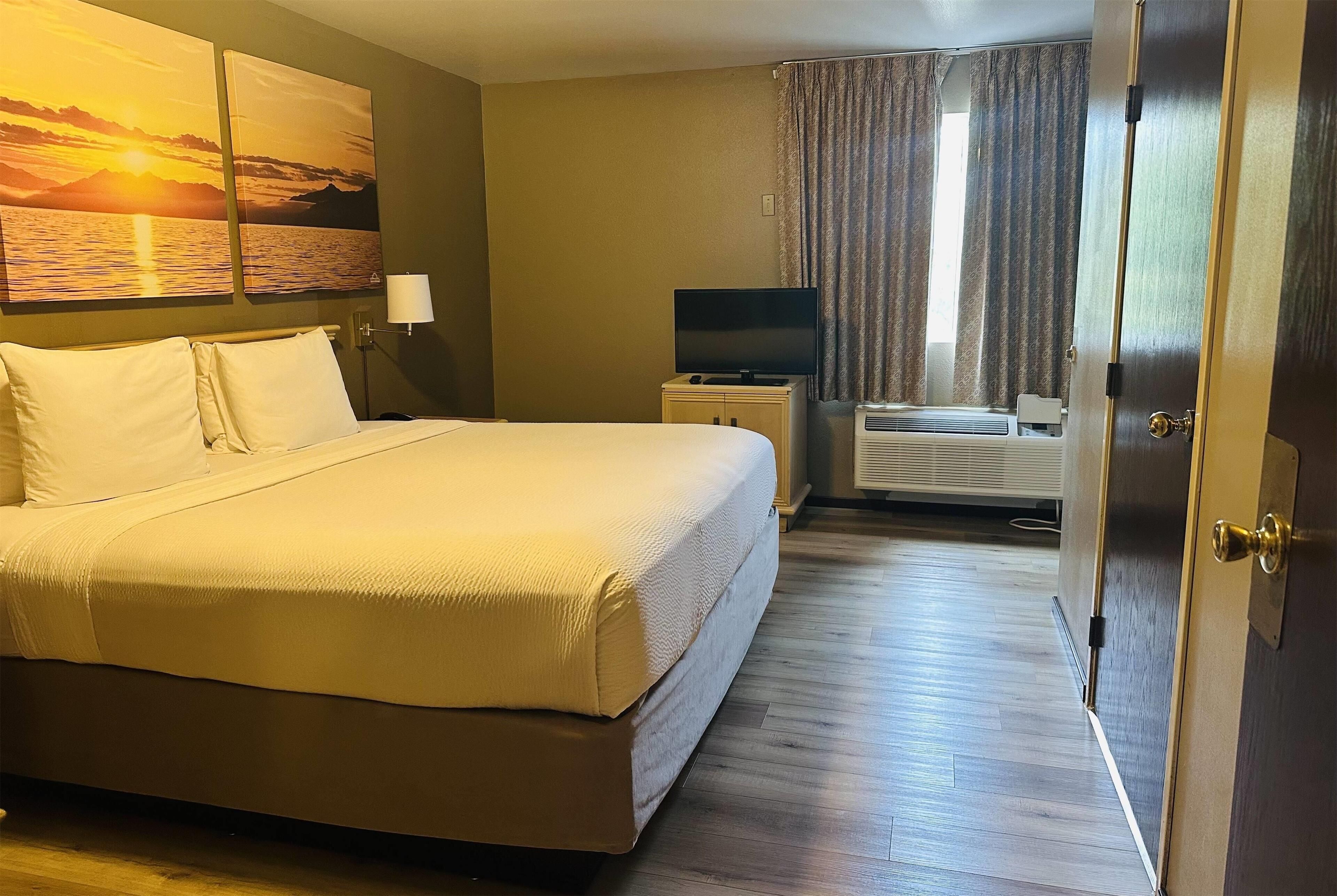 A hotel room with a large bed, a flat-screen TV, an air conditioning unit, and a painting of a sunset over water on the wall.