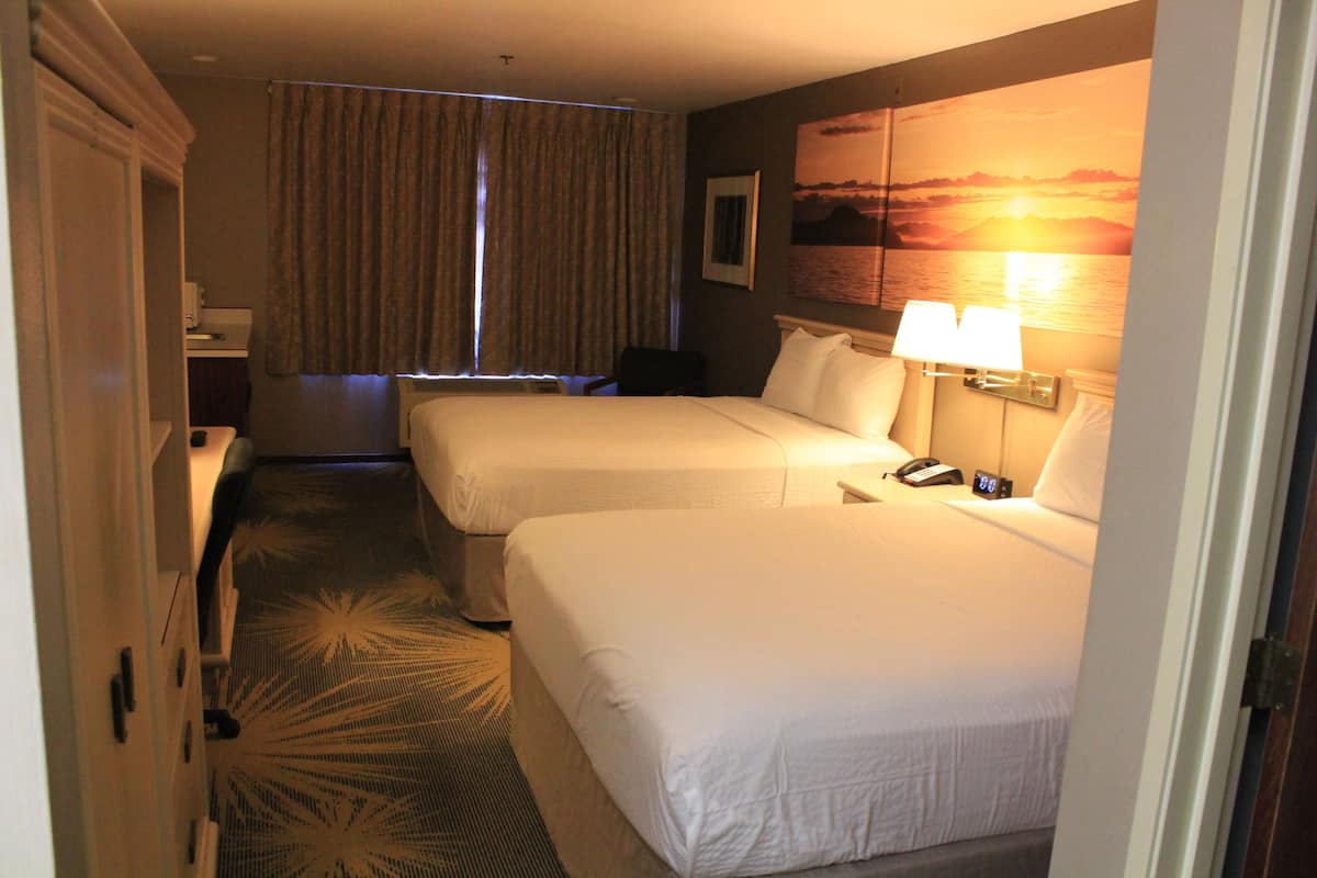 A hotel room with two beds, a desk, a chair, a nightstand, a lamp, and a painting of a sunset.
