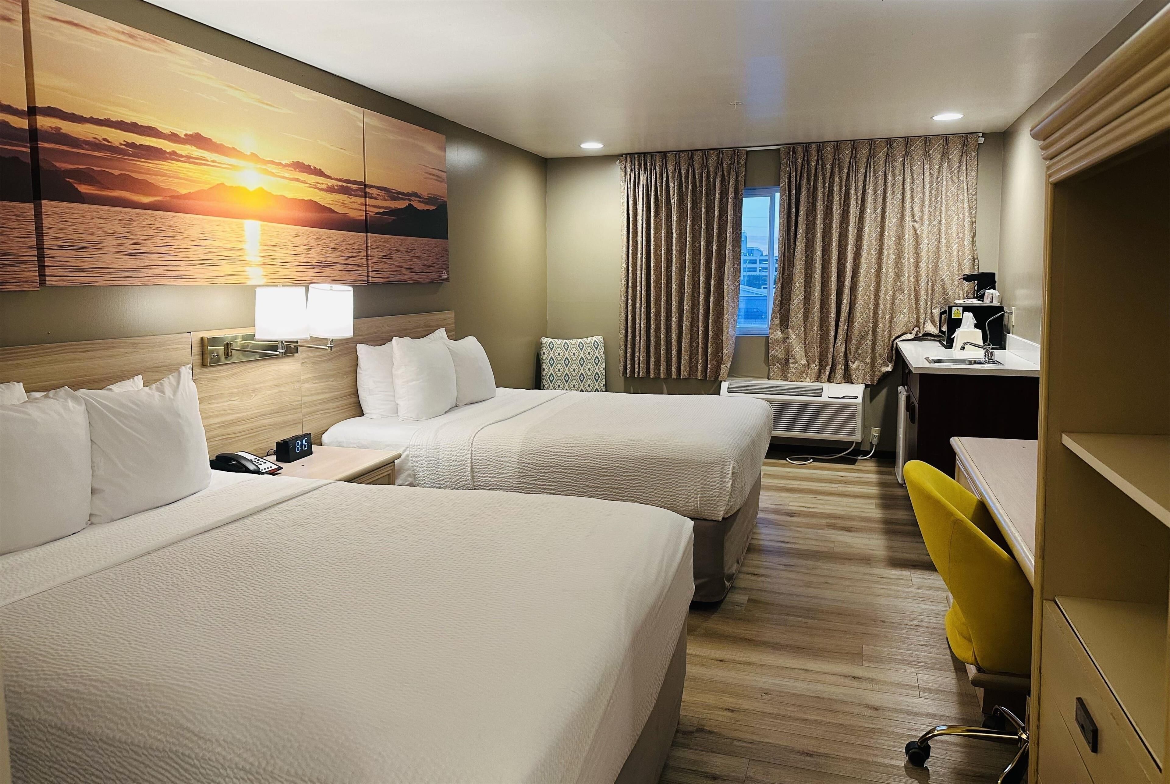 Habitación de hotel con dos camas, un escritorio con silla, una ventana con cortinas, aire acondicionado de pared y un cuadro de un atardecer.