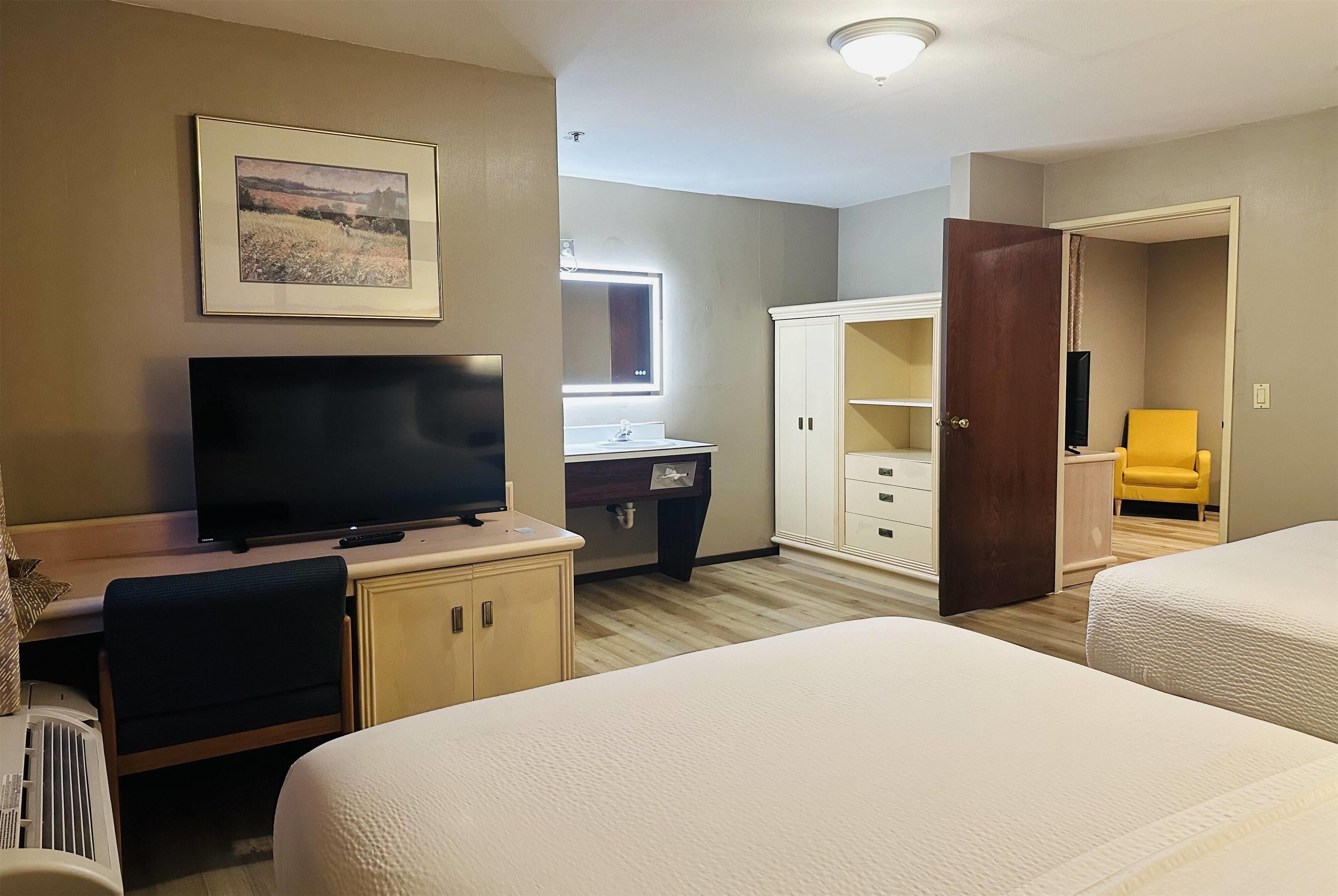 Habitación de hotel con dos camas, un escritorio con televisor, baño con espejo y una silla amarilla.