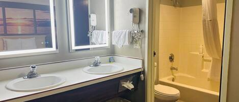 Une salle de bain d’hôtel avec deux lavabos, un miroir, un sèche-cheveux, un rideau de douche et des toilettes.