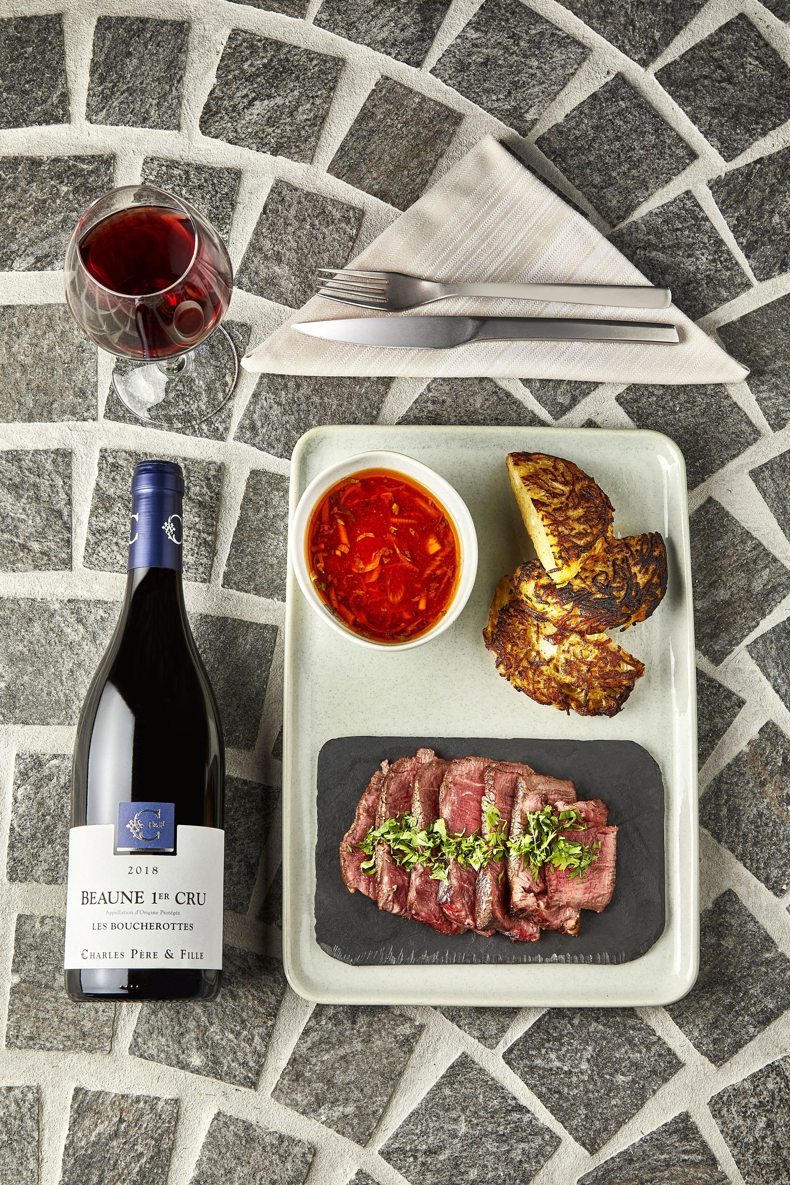 A bottle of Beaune 1er Cru wine, a plate with steak, grilled potatoes, and a bowl of red sauce, all on a tiled surface.