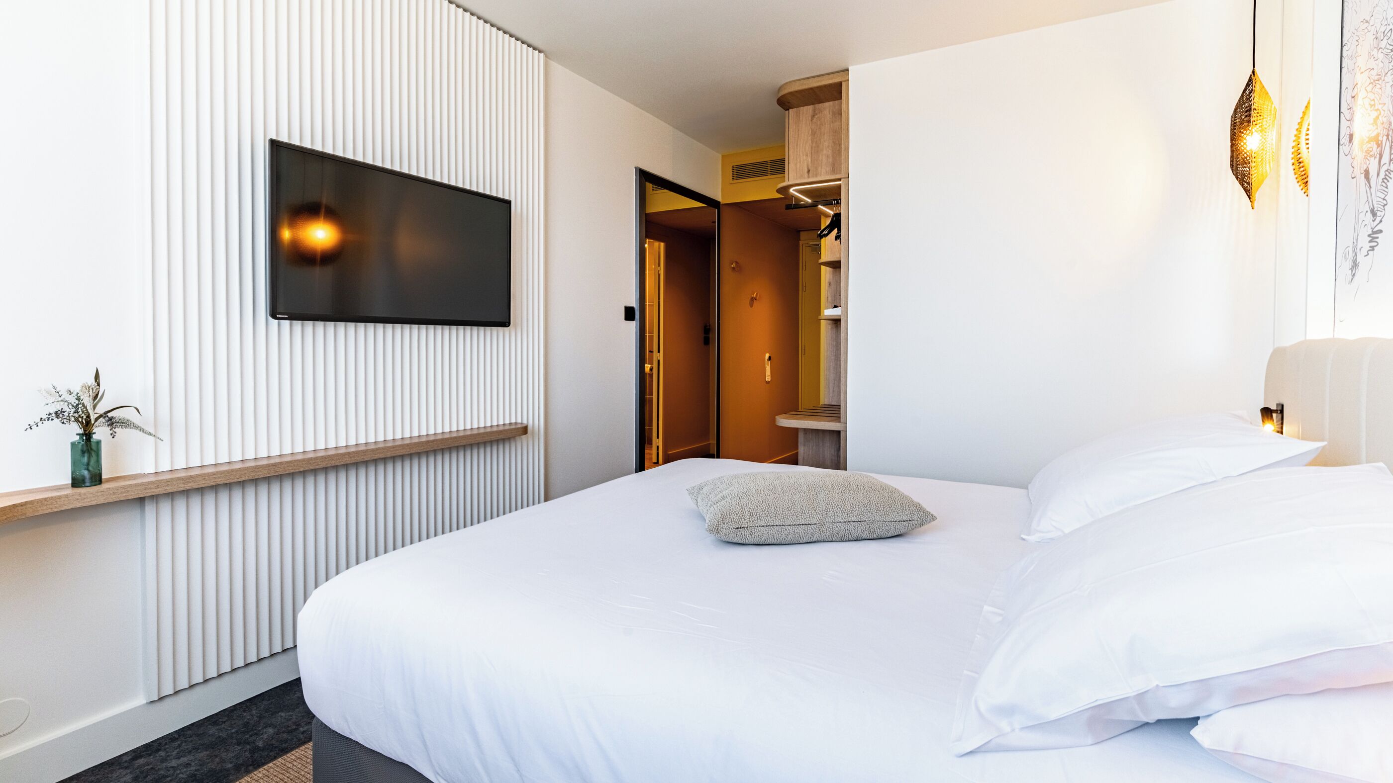 Una habitación de hotel moderna con una cama grande, un televisor de pantalla plana montado en la pared y una mesita de noche con un jarrón de flores.