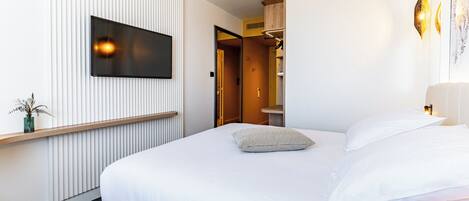 A modern hotel room with a large bed, a flat-screen TV mounted on the wall, and a bedside table with a vase of flowers.