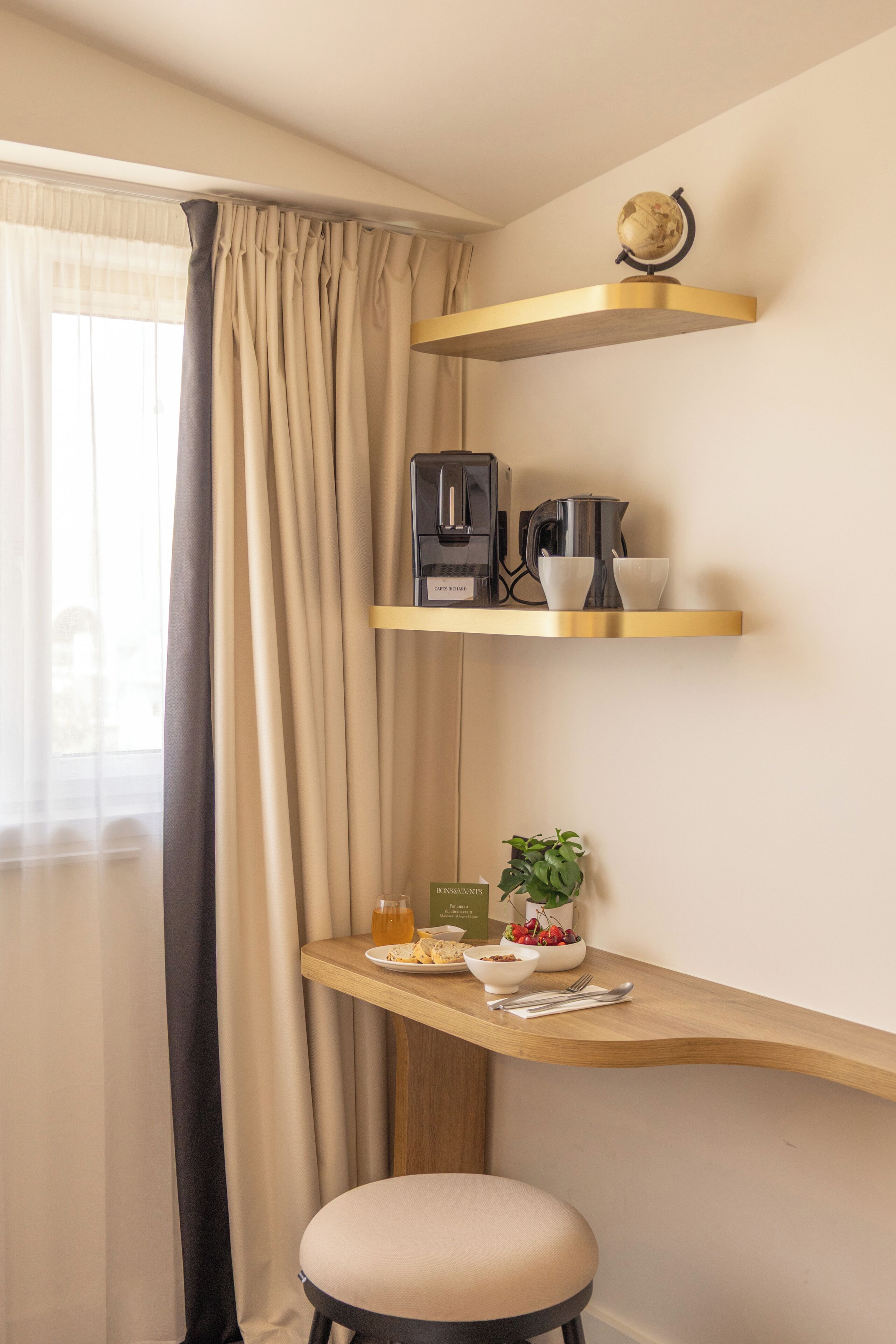A kitchenette with a coffee maker, cups, a bowl of fruit, and a plant on a wooden shelf.