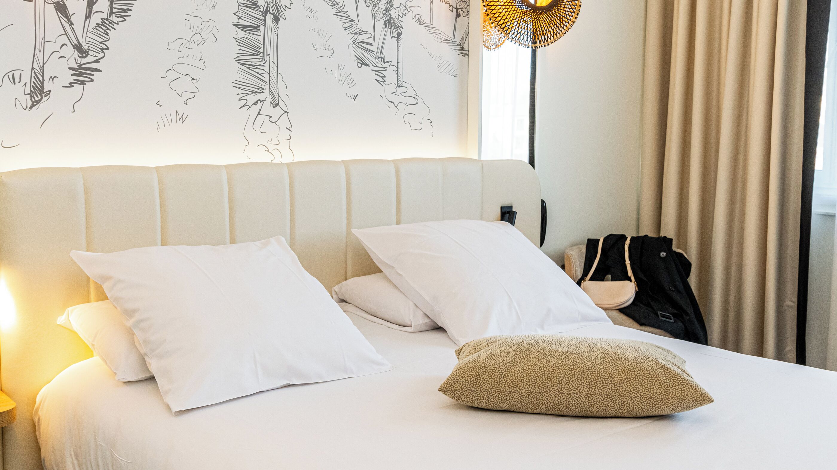 Hotelkamer met een bed, witte kussens, een gestructureerd kussen, een lamp, een kapstok en een raam met gordijnen.