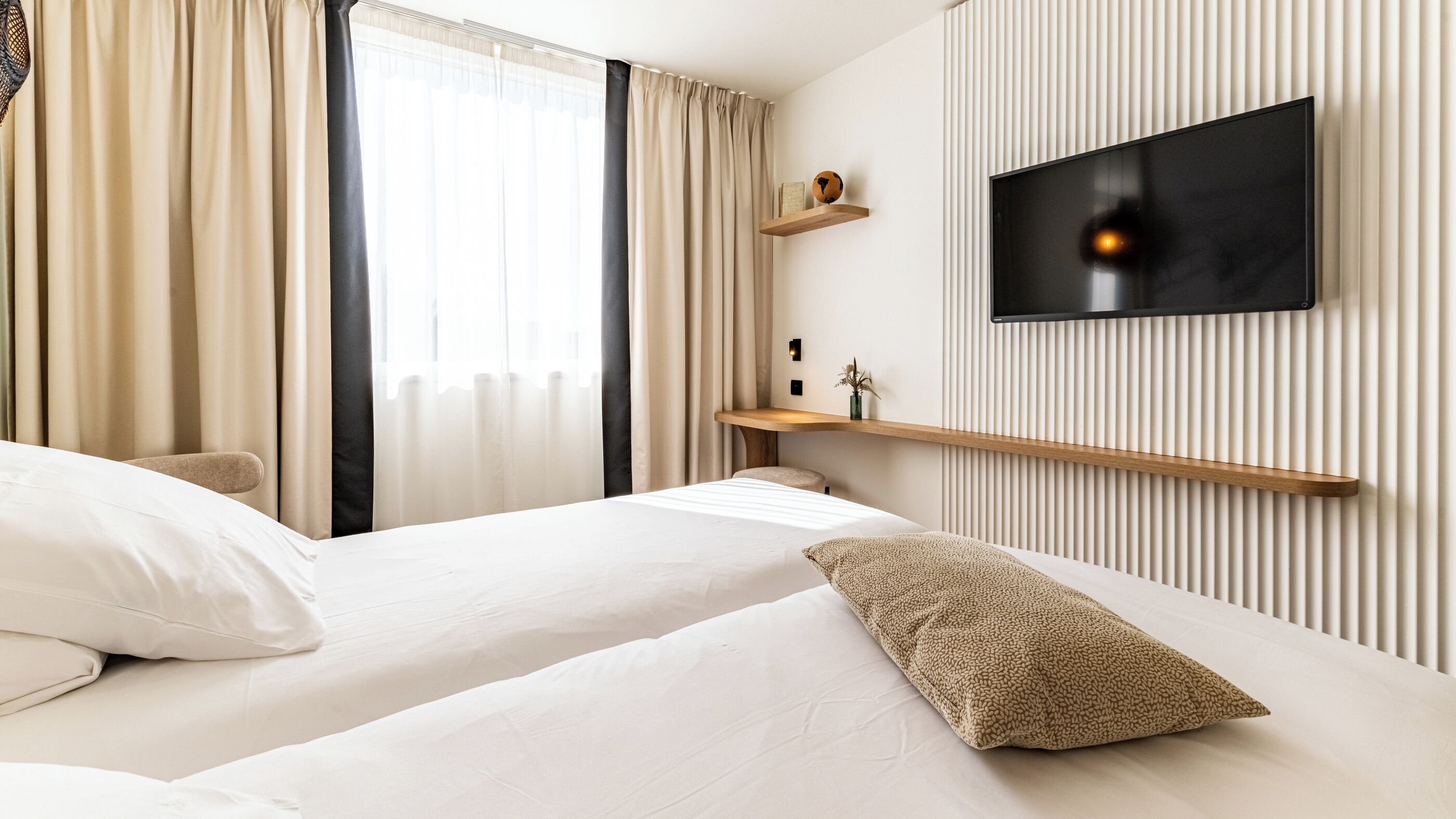 Een hotelkamer met twee bedden, een aan de muur bevestigde televisie en een raam met gordijnen.