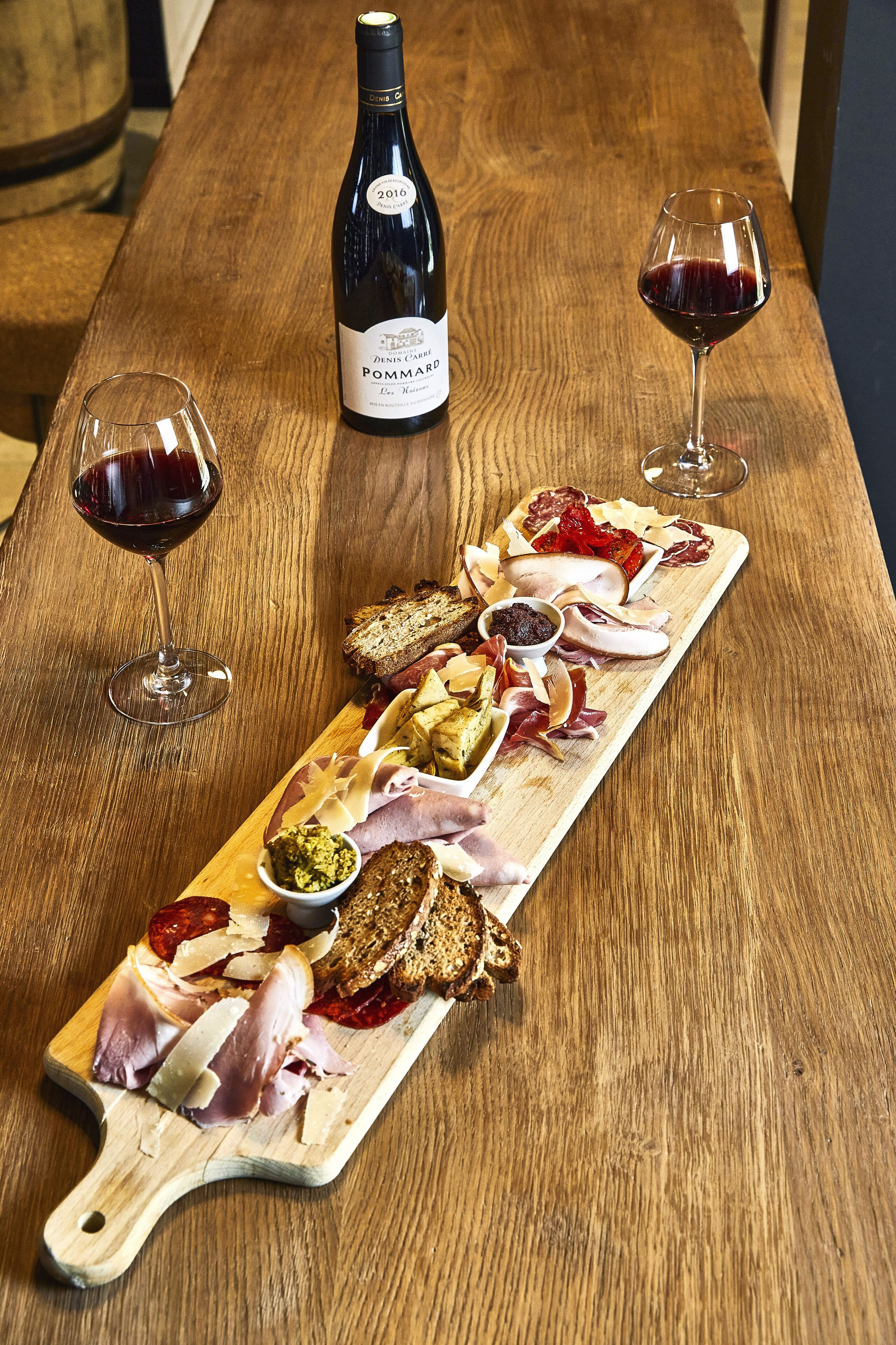 Ein Holztisch mit einer Weinflasche, zwei Weingläsern und einer Wurstplatte mit verschiedenen Fleisch-, Käse- und Brotsorten.