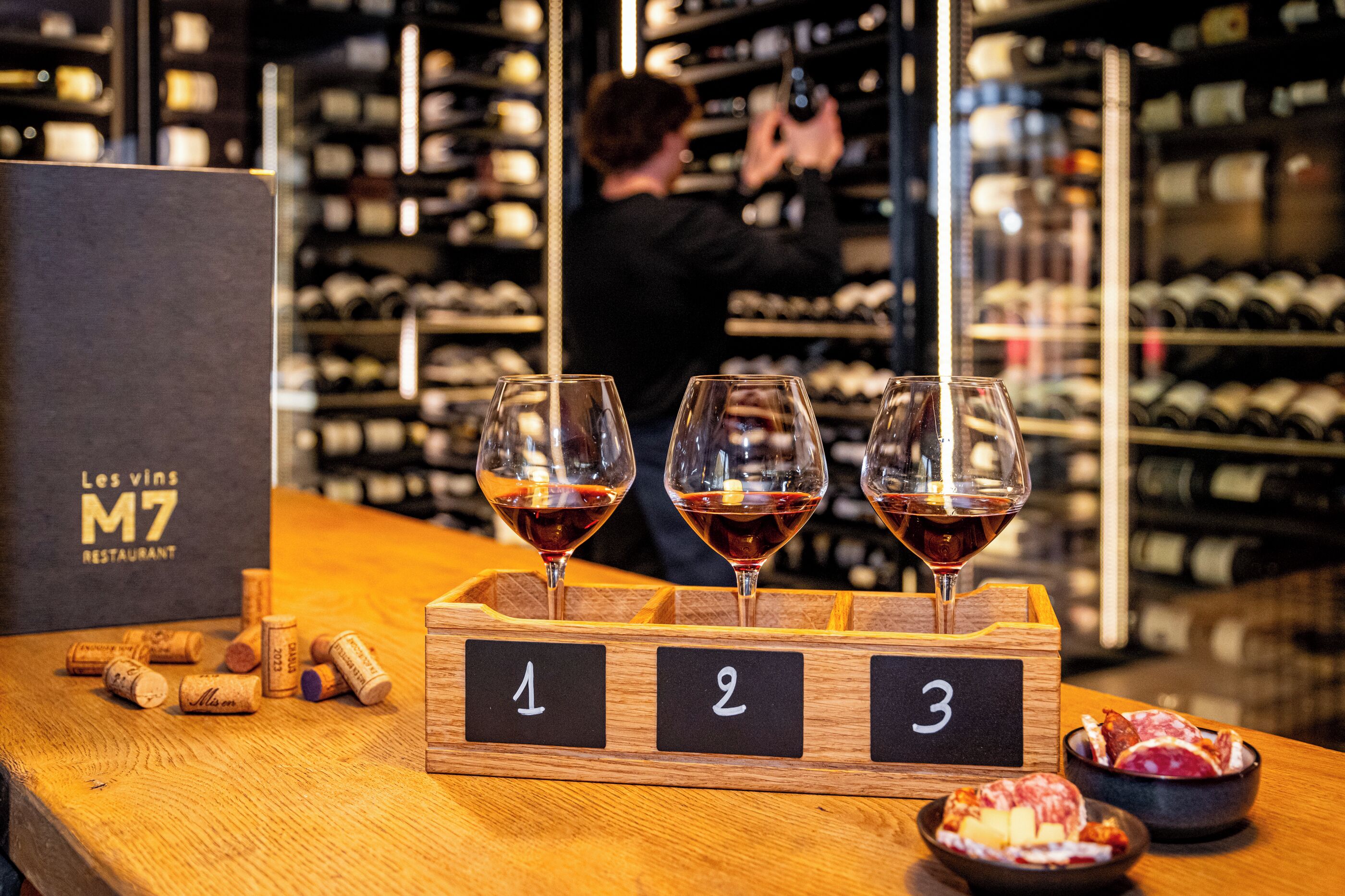 Viininmaistelutilaisuus, jossa on kolme lasillista punaviiniä puisen jalustan päällä. Lasit on numeroitu 1, 2 ja 3. Taustalla on laatikko, jossa lukee: ”Les vins M7 -ravintola”.