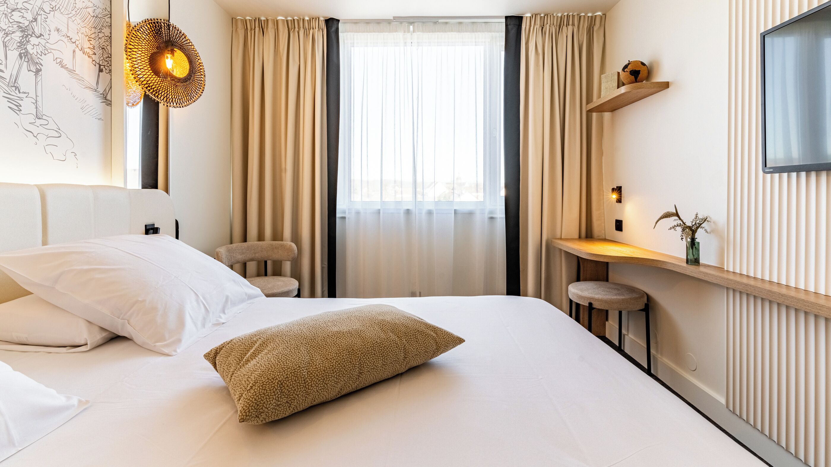 A hotel room with a large bed, a small desk, a chair, a wall-mounted shelf, and a decorative wall hanging.