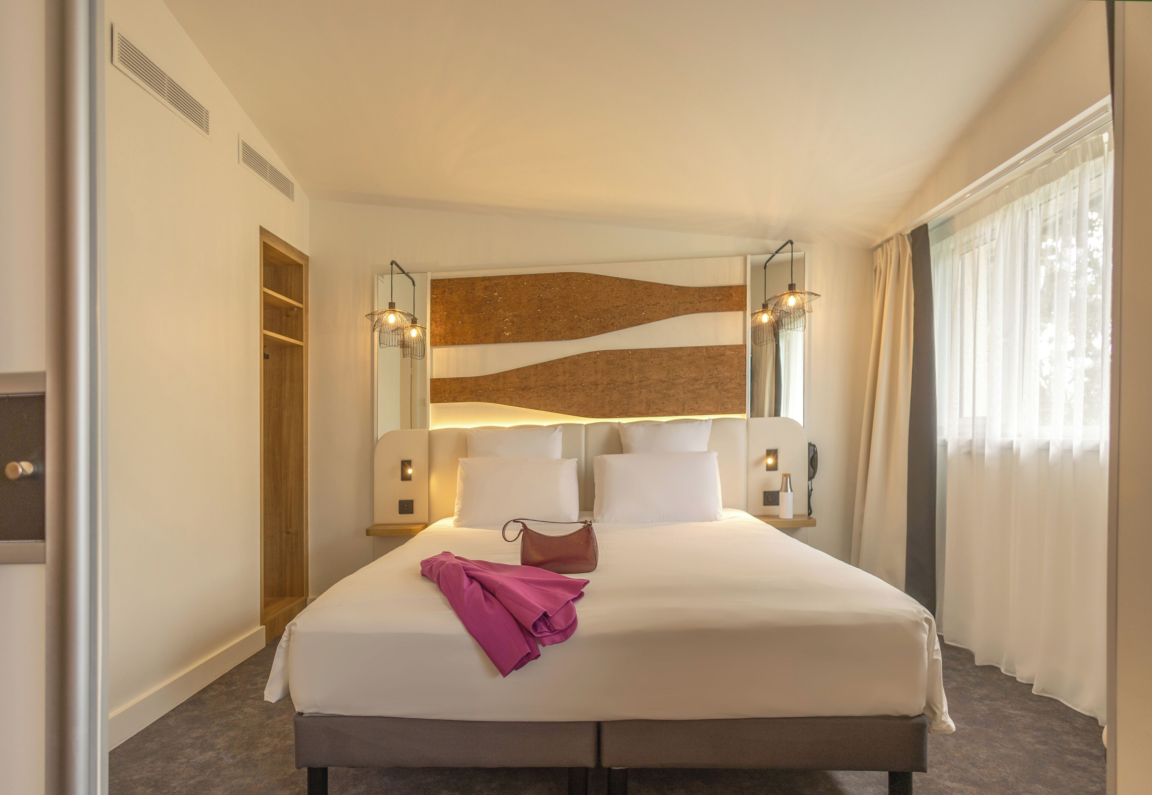 A hotel room with a large bed, a wooden headboard, a nightstand, a lamp, and a window with curtains.