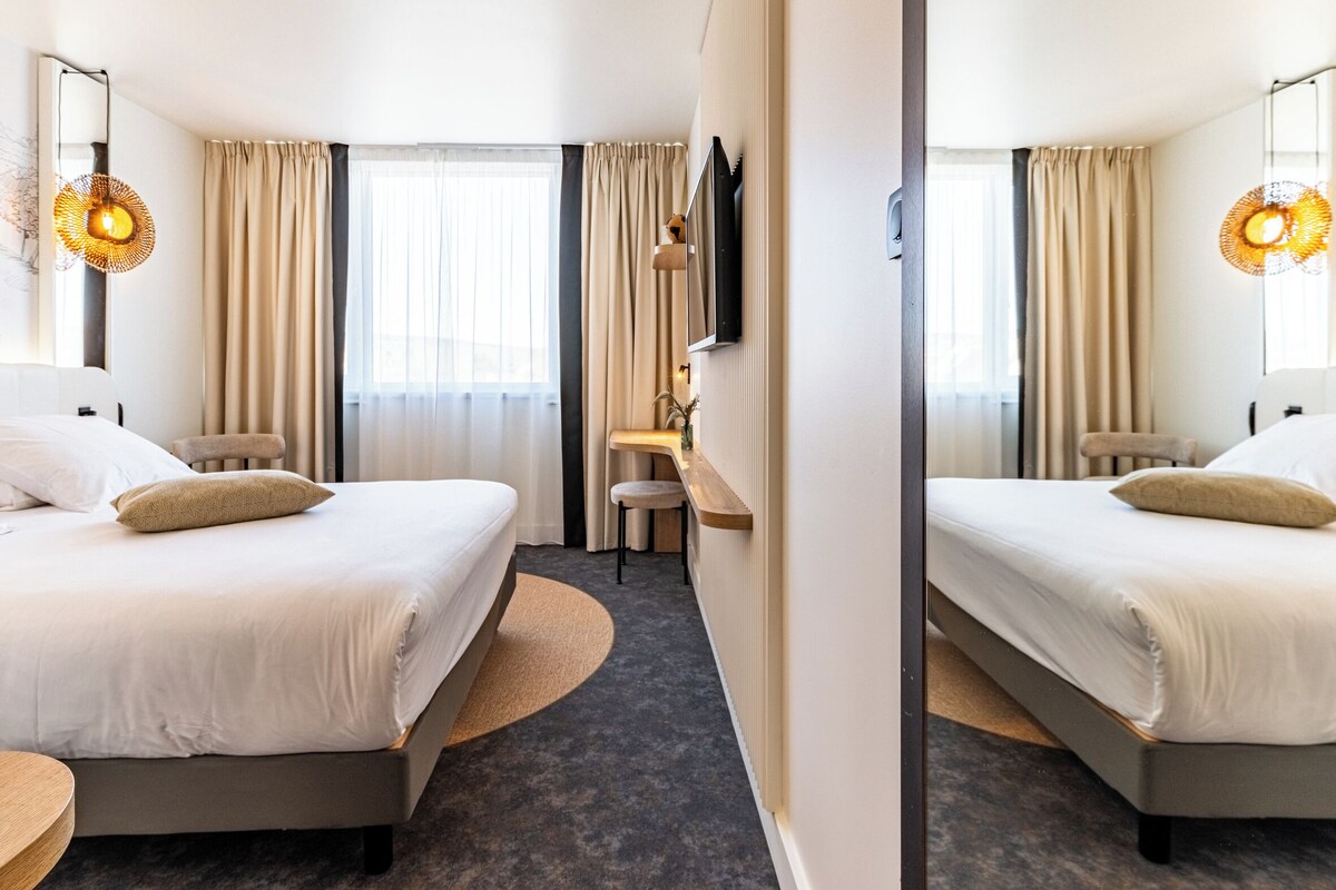 A hotel room with two beds, a desk, a chair, a TV, and a large window with curtains.