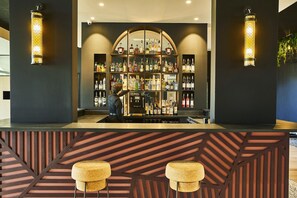 A bar area with a bartender, a well-stocked liquor shelf, and two stools. - Mercure Beaune Centre (Beaune)