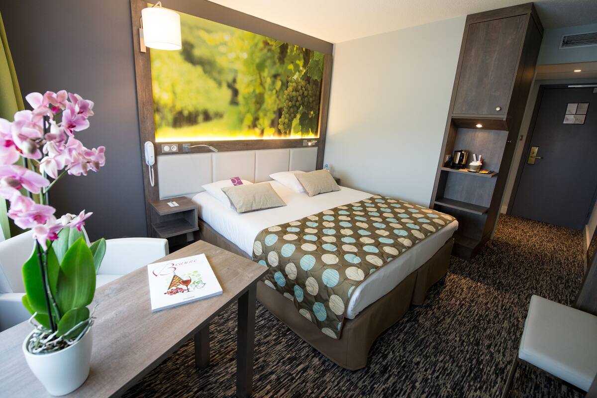 A hotel room with a bed, bedside tables, a desk with a plant, a painting on the wall, and a wardrobe.