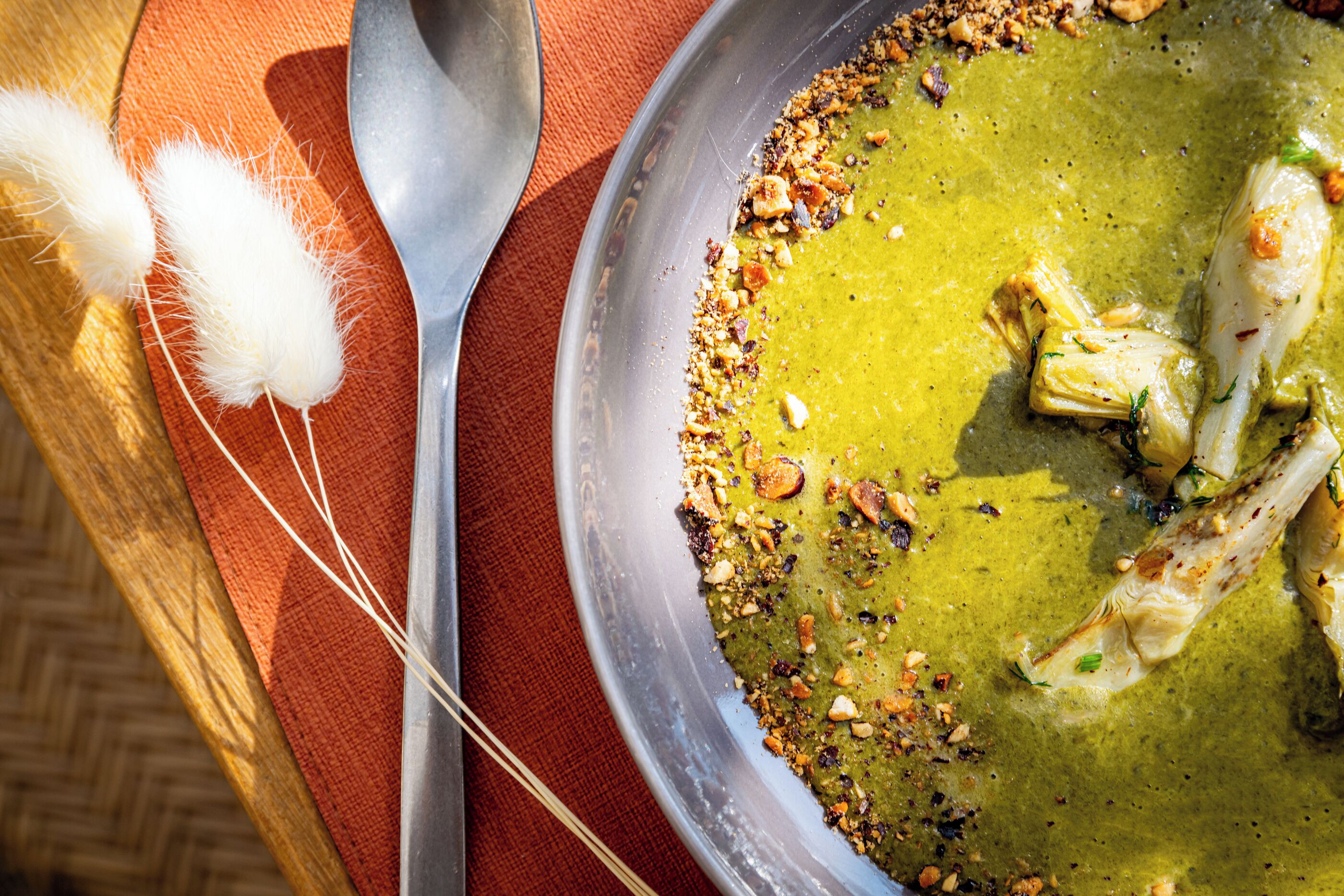 A bowl of green soup with nuts and herbs, a spoon, and a napkin with a fork on a wooden table.